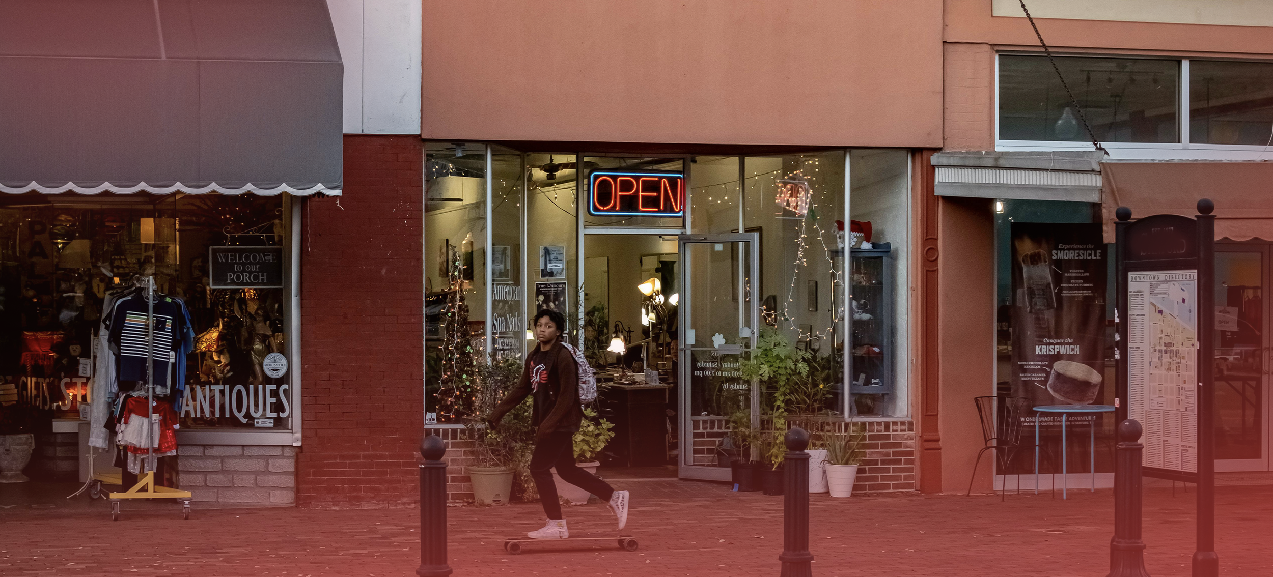 A storefront with a neon 'Open' sign, glass doors, and windows displaying various items inside. A young person with a skateboard walks past in front of the shop. To the left, there is an antique store with a window showcasing clothing and a sign that says 'Antiques'. To the right, another shop with a menu board and a small outdoor table. The sidewalk is paved with red bricks, and there are black posts along the street.