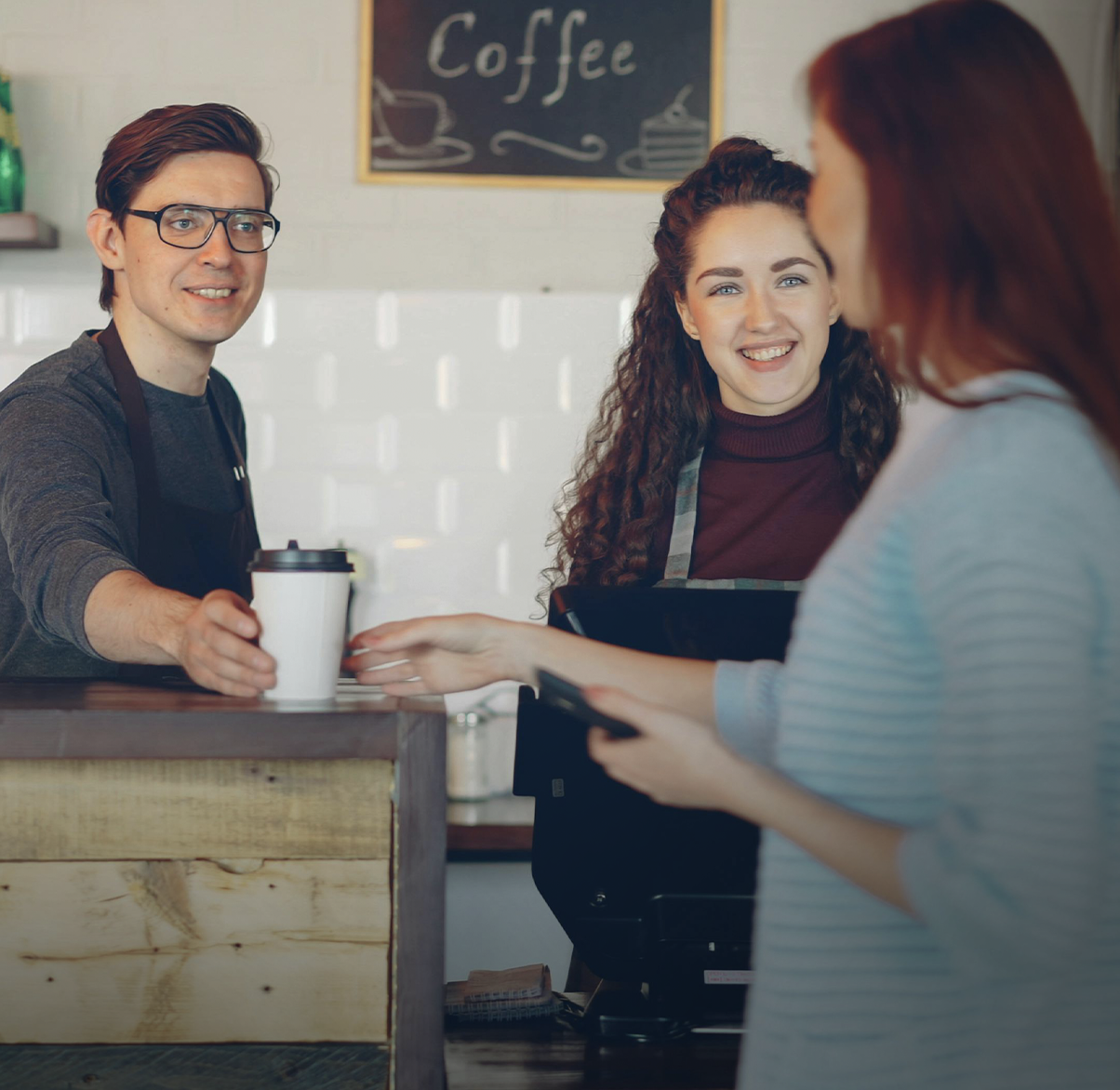 A barista handing a coffee to a customer while another customer waits and smiles at the counter in a coffee shop.