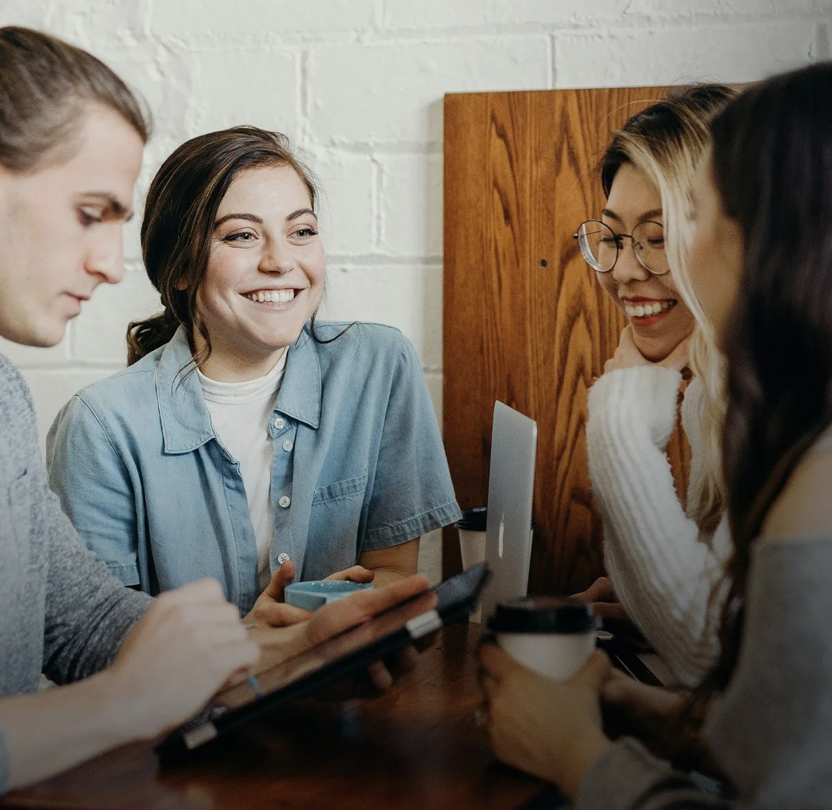 Four young women sitting around a wooden table, smiling and talking in a cozy indoor setting with a white brick wall and a wooden panel behind them.