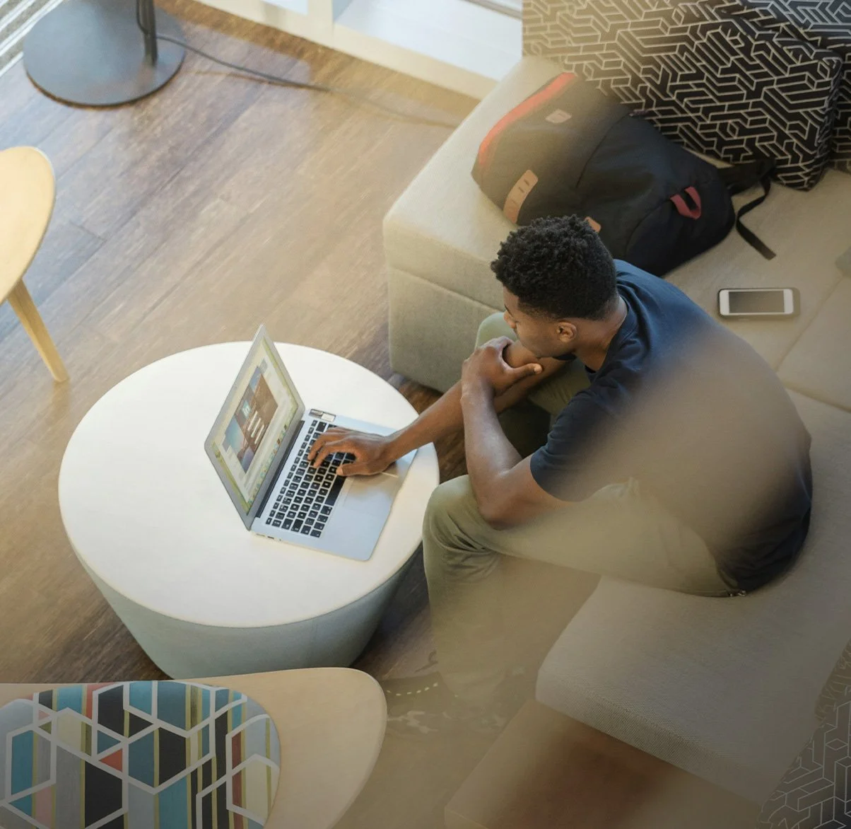 A man sitting on a beige couch using a laptop in a living room, with a white round coffee table in front of him, a black backpack on the couch, and a smartphone on the couch armrest.