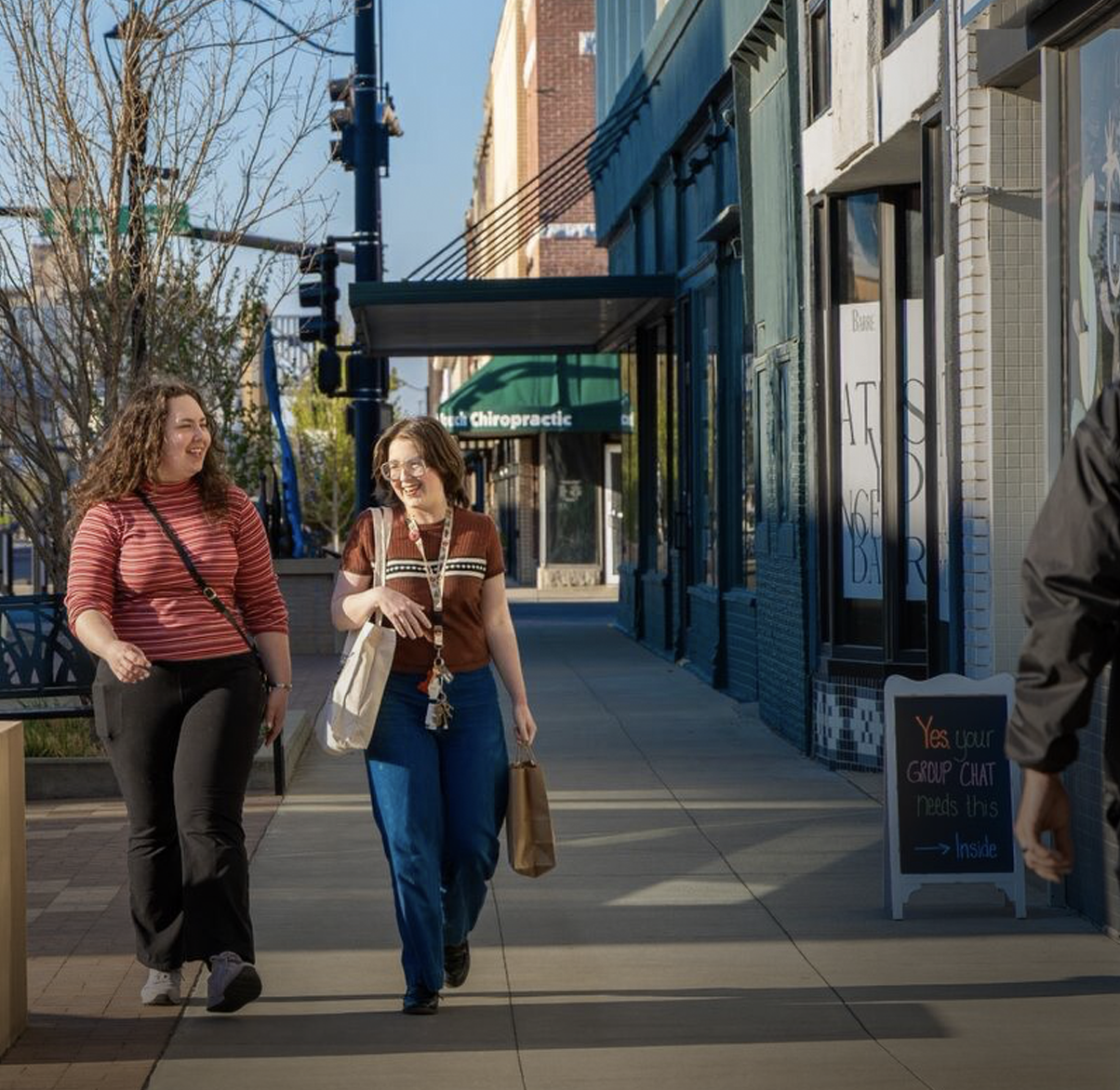 Two women walking and talking on a sunny sidewalk in an urban area, smiling and holding shopping bags.