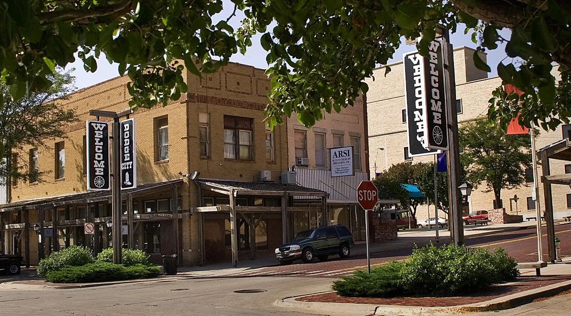 Downtown street corner with brick buildings, signs reading "Welcome Dodge City" and "Visit Enigma"; stop sign, parked cars, trees, and bushes.