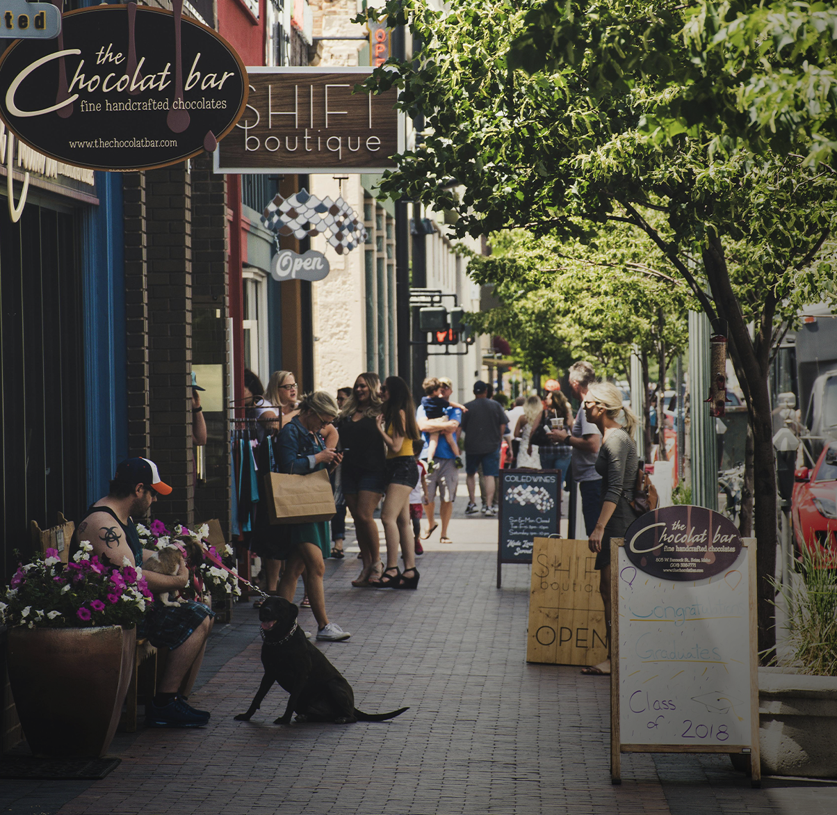 People walking and standing on a busy sidewalk outside shops, including a chocolate shop with signs, some trees, and a man sitting on a bench with a dog.