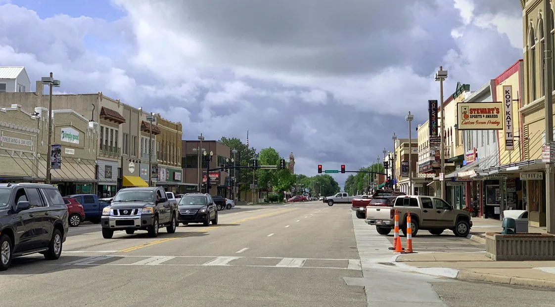 Main street in a small town with parked cars along both sides, storefronts on the right, traffic lights in the distance, cloudy sky overhead.