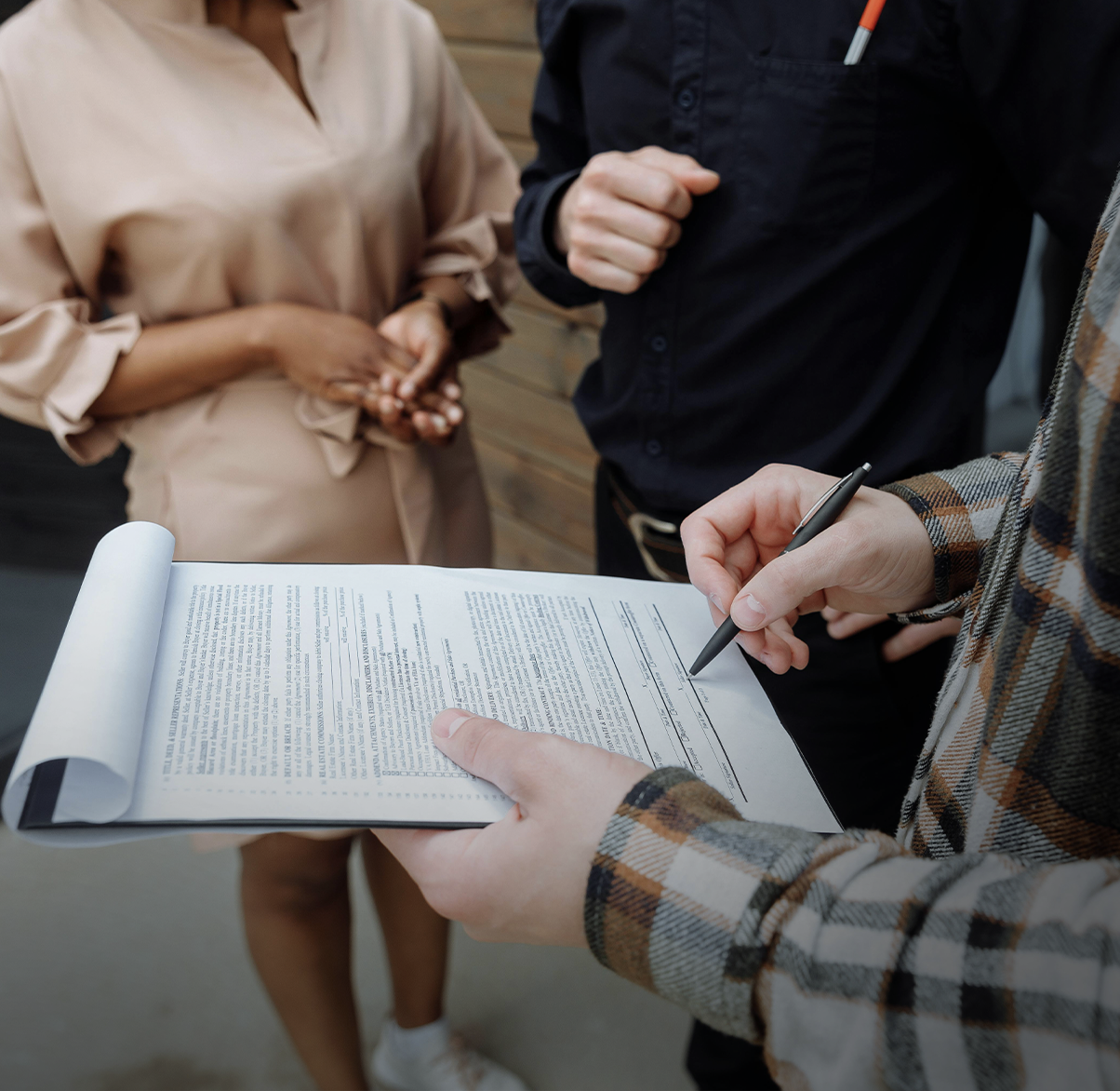 Person signing a legal document with a pen while two others stand nearby, holding hands and waiting.