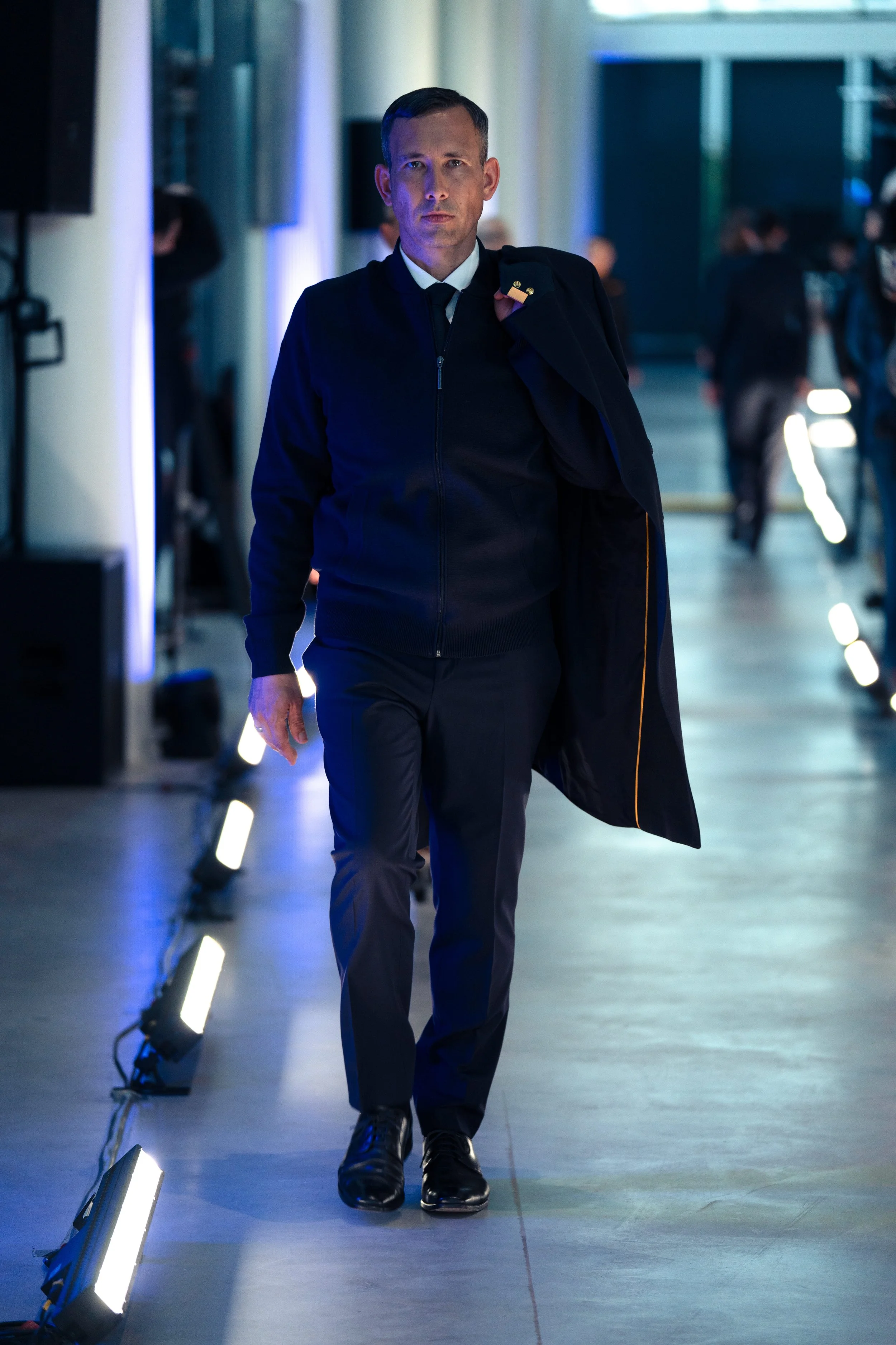 Bild von dem Catwalk des Release-Events der neuen Lufthansa Dienstkleidung 2026: Ein Mann in einem schwarzen Anzug läuft in einem modernen Gebäude, während er eine Jacke über die Schulter trägt.