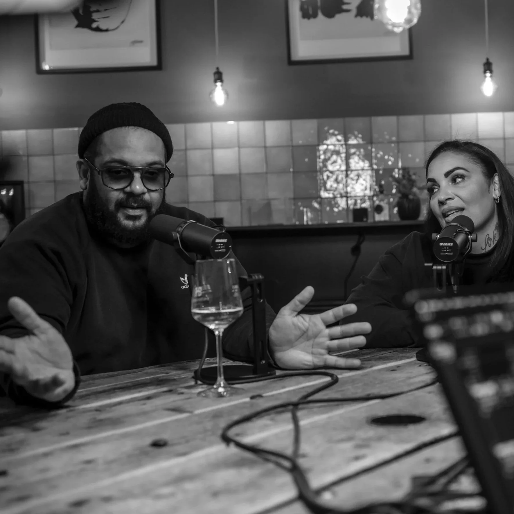 Momentaufnahme aus dem Podcast Roomgebabbel mit Tiffany Kirkland und Faiz von Bro'Sis: Zwei Menschen sprechen während eines Podcasts, in einem Raum mit moderner Inneneinrichtung, Kerzen und Wandkunst, in Schwarz-Weiß-Fotografie.