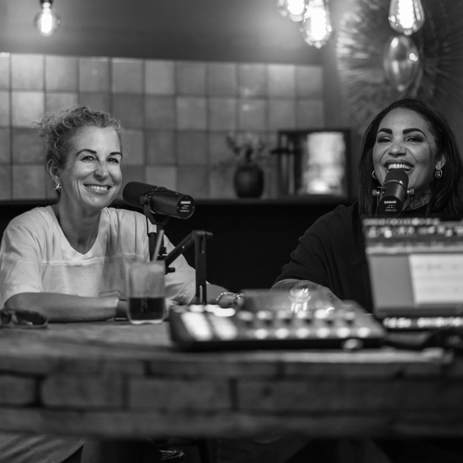 Momentaufnahme aus dem Podcast Roomgebabbel mit Tiffany Kirkland und Daniela Georgi von der CDU Wiesbaden: Zwei Frauen sitzen an einem Tisch, sprechen in Mikrofone und lachen. Im Hintergrund sind Lampen und eine Wand mit Fachwerk zu sehen.