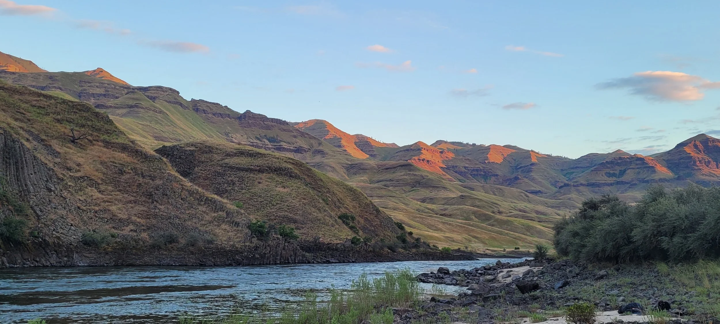 Scenic view of a river flowing through a valley with green hills and mountains under a partly cloudy sky.