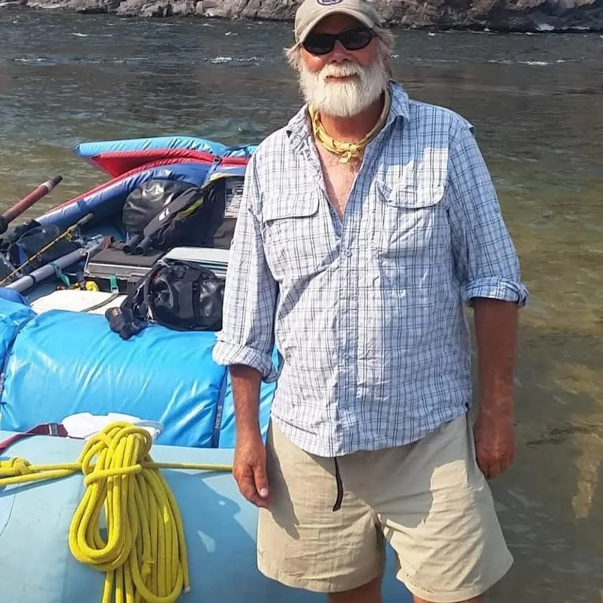 An older man with a white beard, wearing sunglasses, a beige cap, a plaid long sleeve shirt, and beige shorts, standing next to a boat on a river.