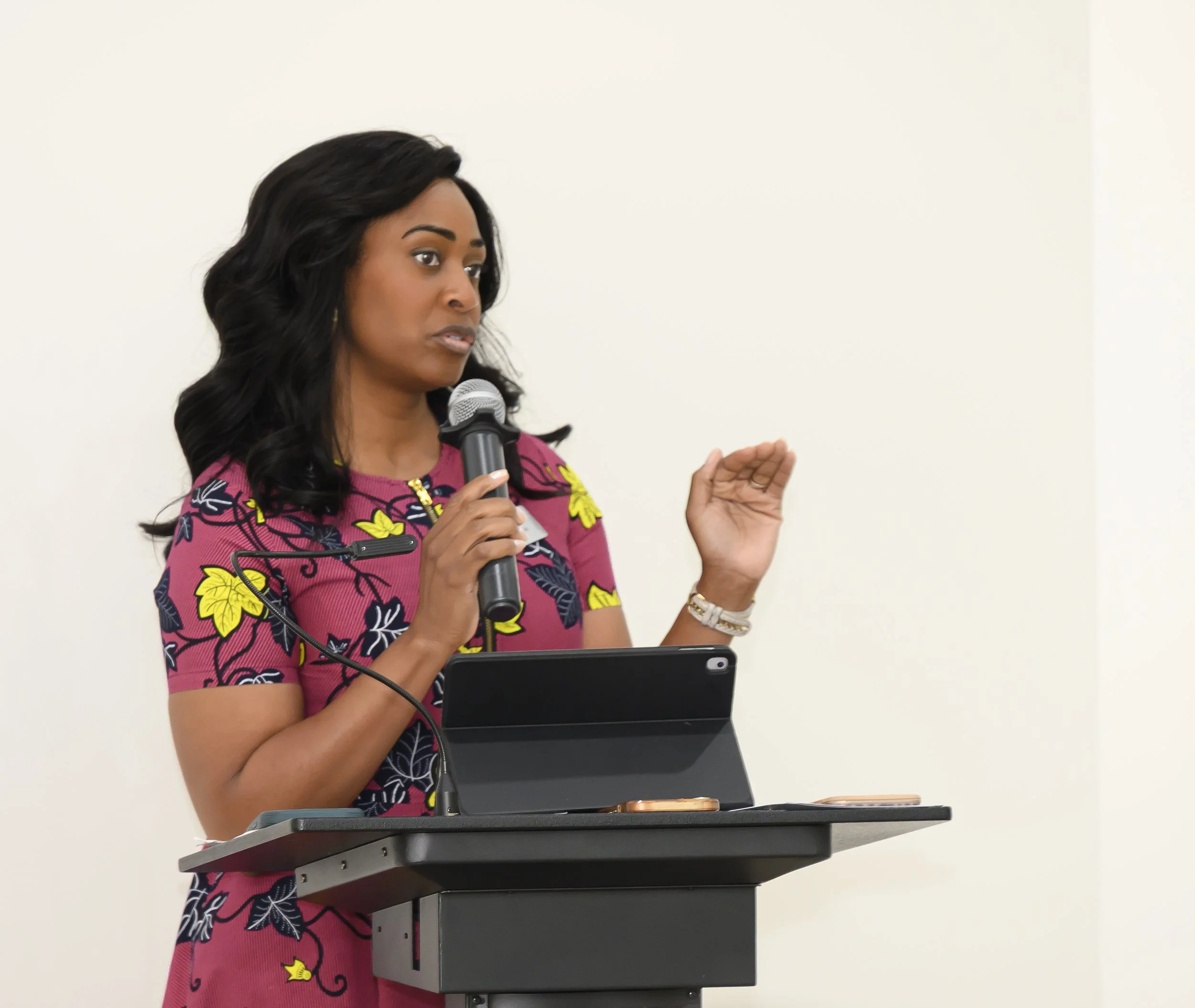 Woman with dark hair wearing a colorful patterned dress speaking into a microphone and gesturing with her hand behind a lectern.