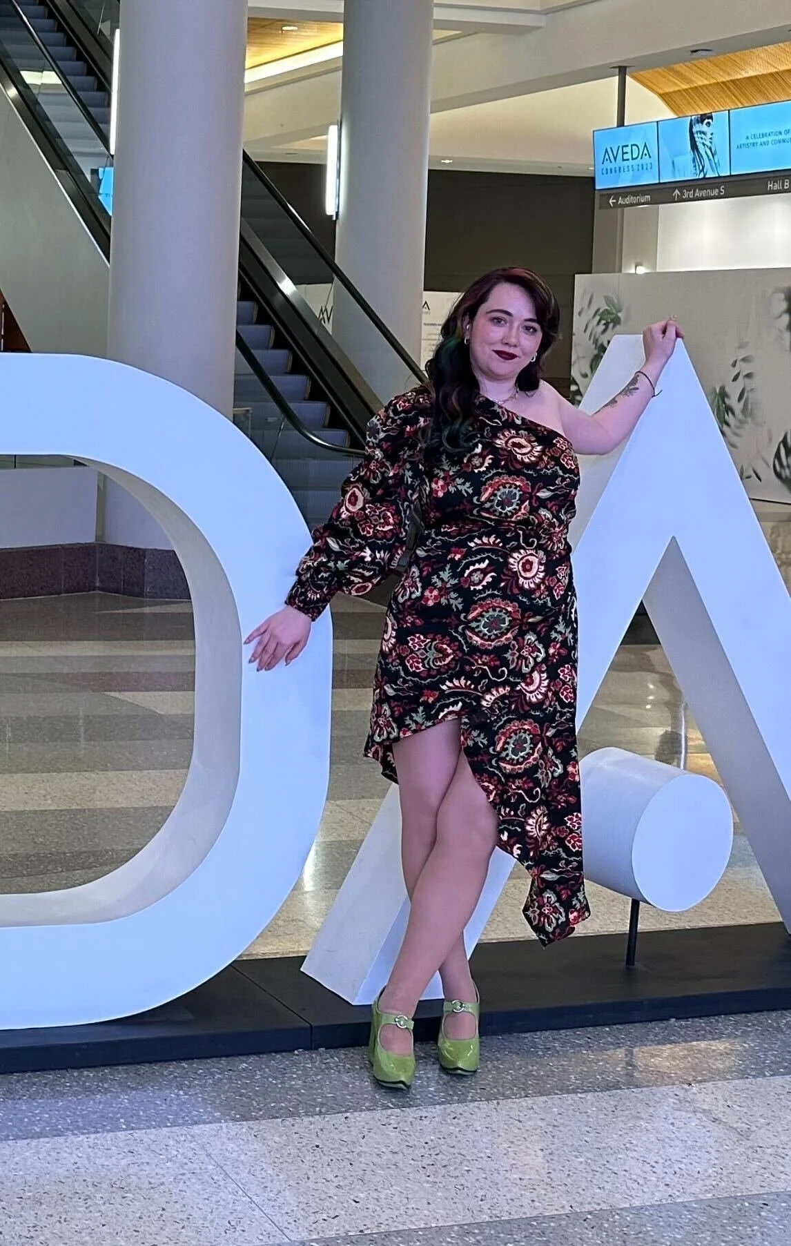 A woman with dark hair and tattoos on her arm standing next to large white 3D letters inside a modern building. The woman is wearing a dark floral dress and lime green high heels.