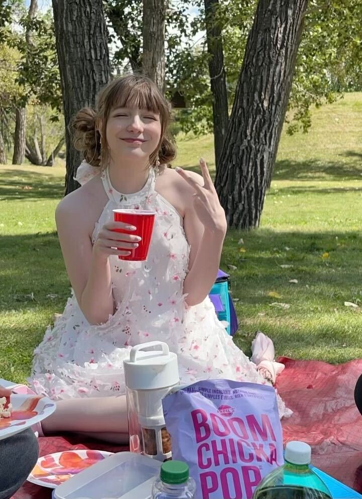 A young woman with short brown hair, wearing a white dress with pink floral patterns, sits on a picnic blanket outdoors, holding a red cup and making a peace sign. There are trees and grass in the background, and picnic items like snacks and drinks in front of her.