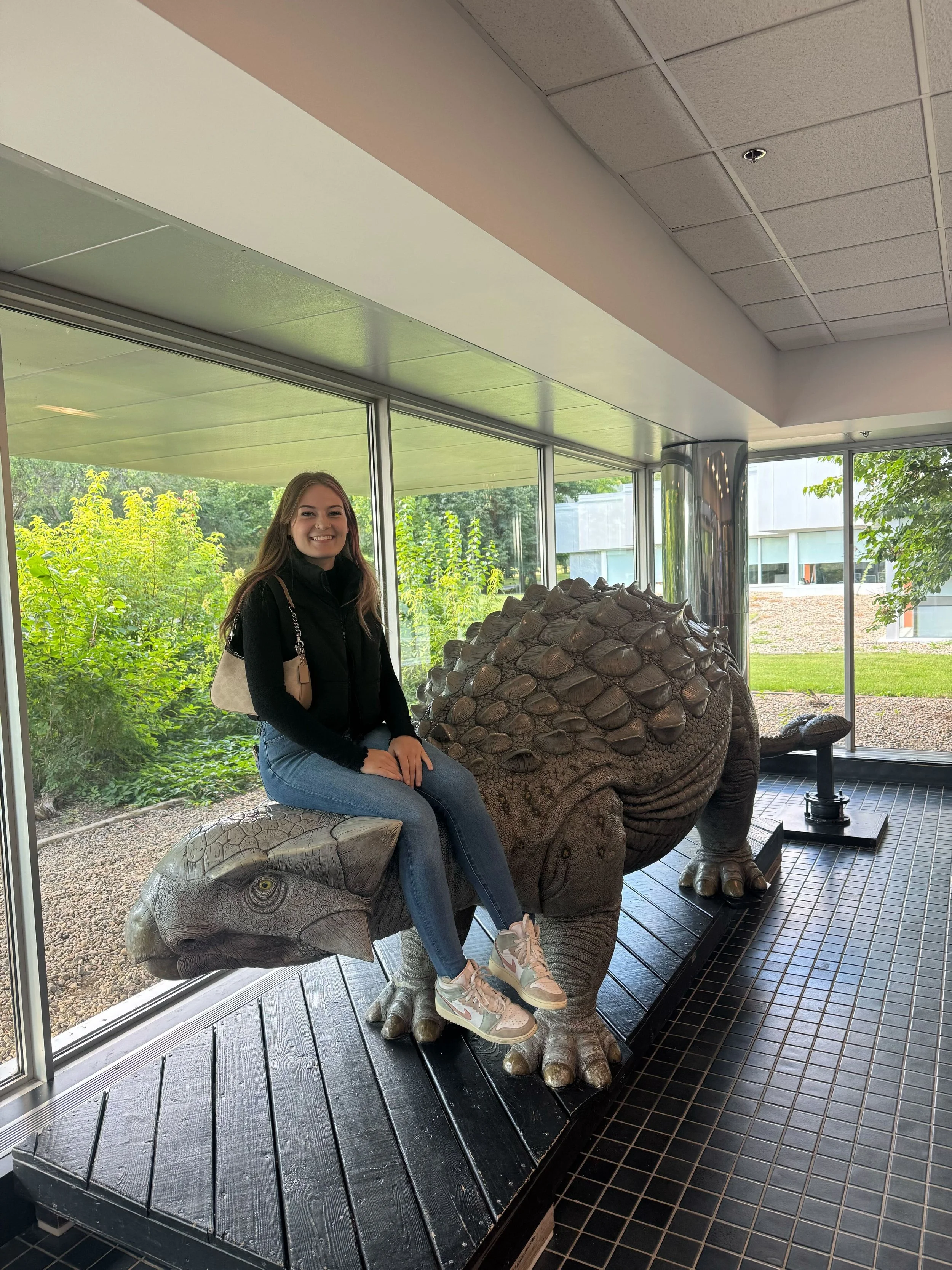 A young woman sitting on a decorative turtle sculpture inside a building with large windows showing greenery outside.
