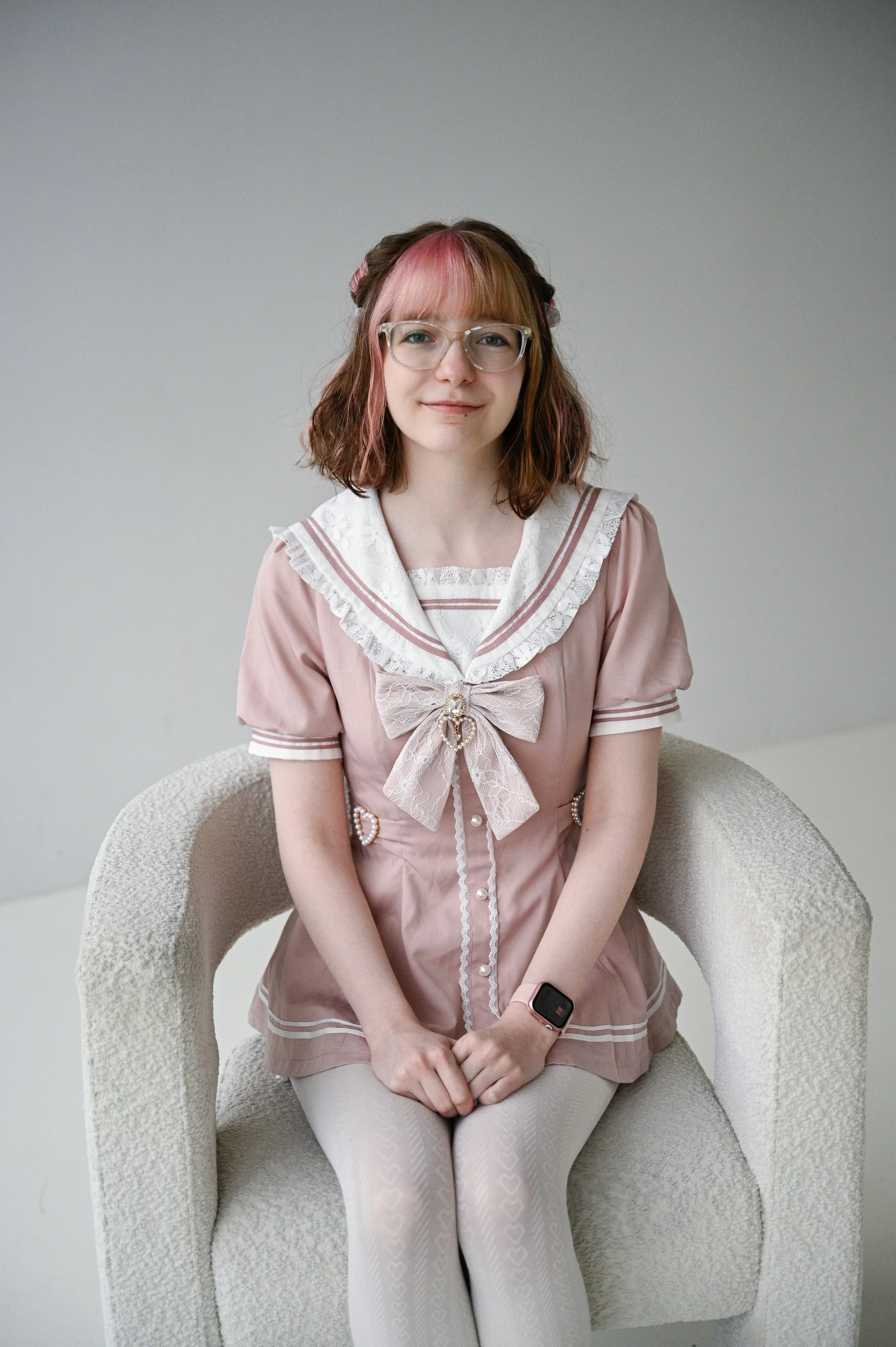 A young woman with pink and brown hair sitting on a beige armchair, wearing glasses, a pink and white vintage-style dress, and a smartwatch.