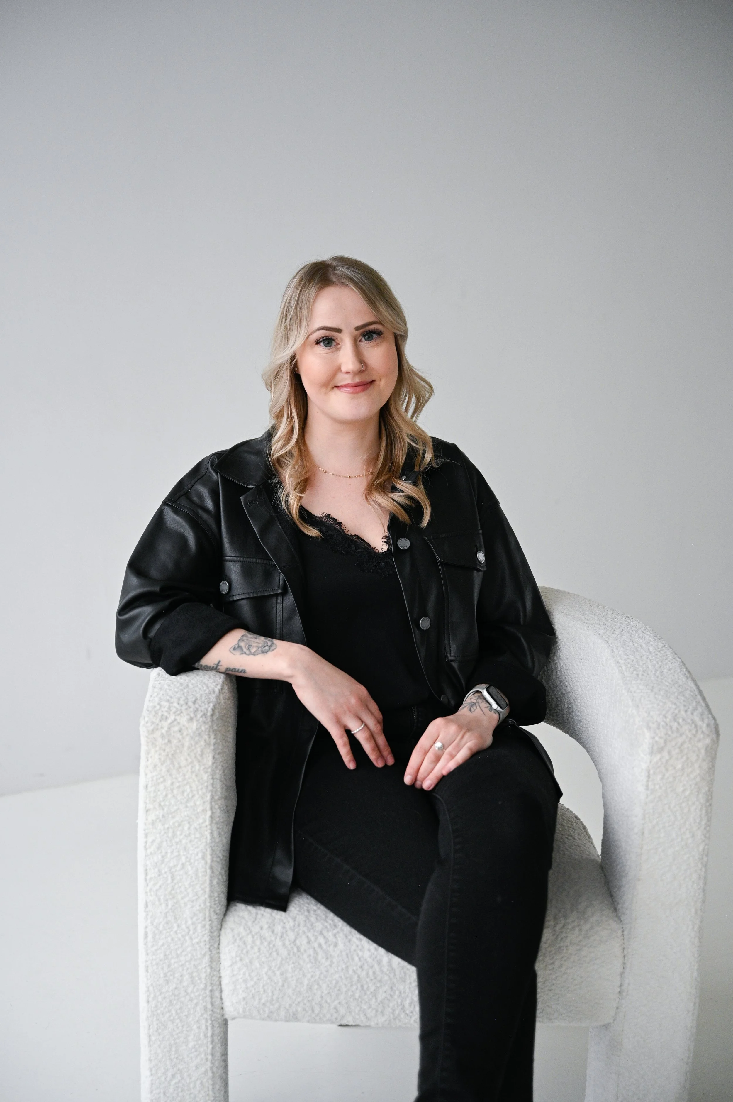 A woman with blonde hair, wearing a black leather jacket and black pants, sitting on a white textured armchair against a light gray wall.