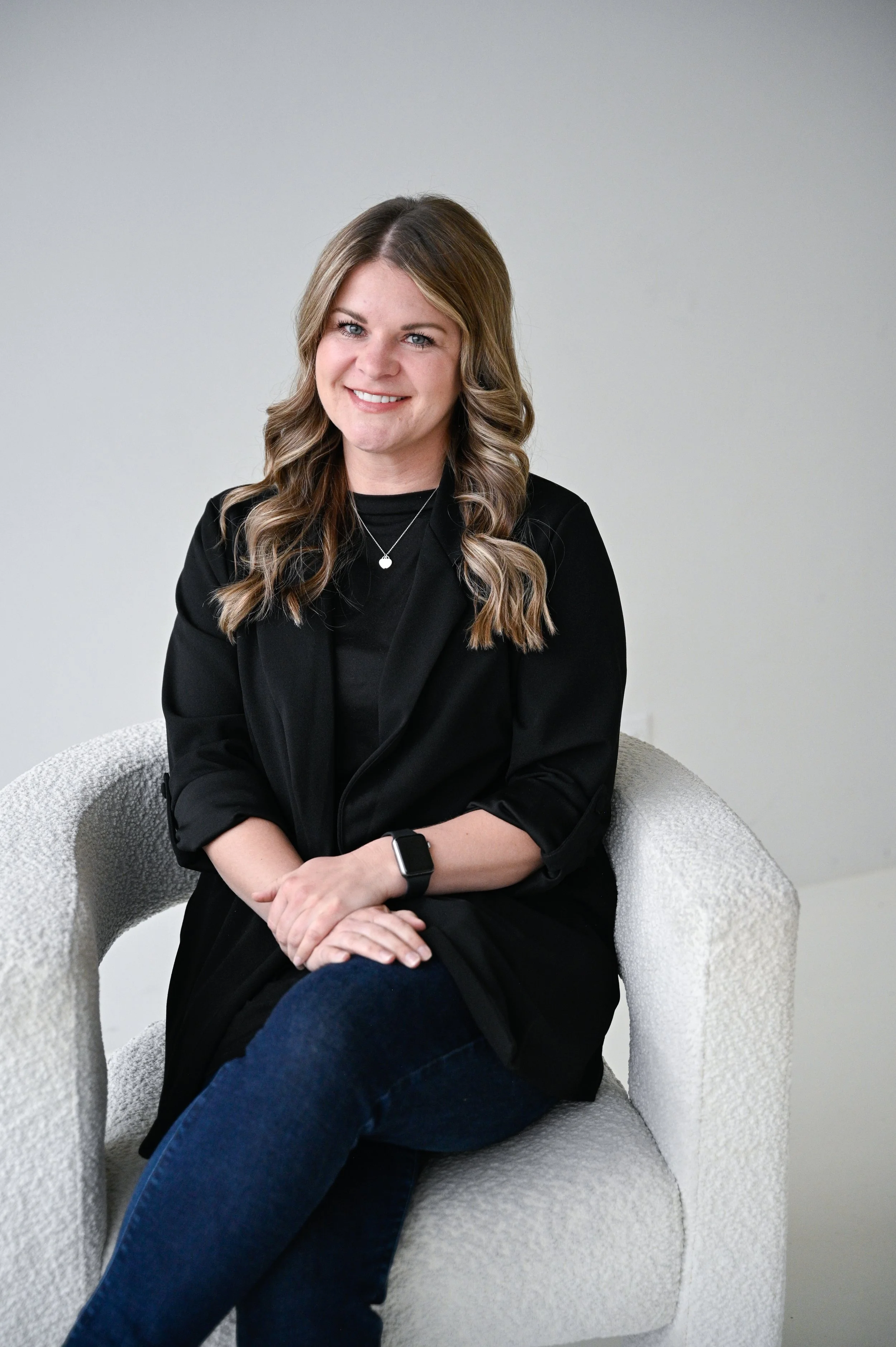 A woman with wavy light brown hair, smiling, sitting on a light-colored textured armchair against a plain white wall. She is wearing a black jacket, black shirt, and blue jeans, with a smartwatch on her left wrist and a necklace with a small pendant.