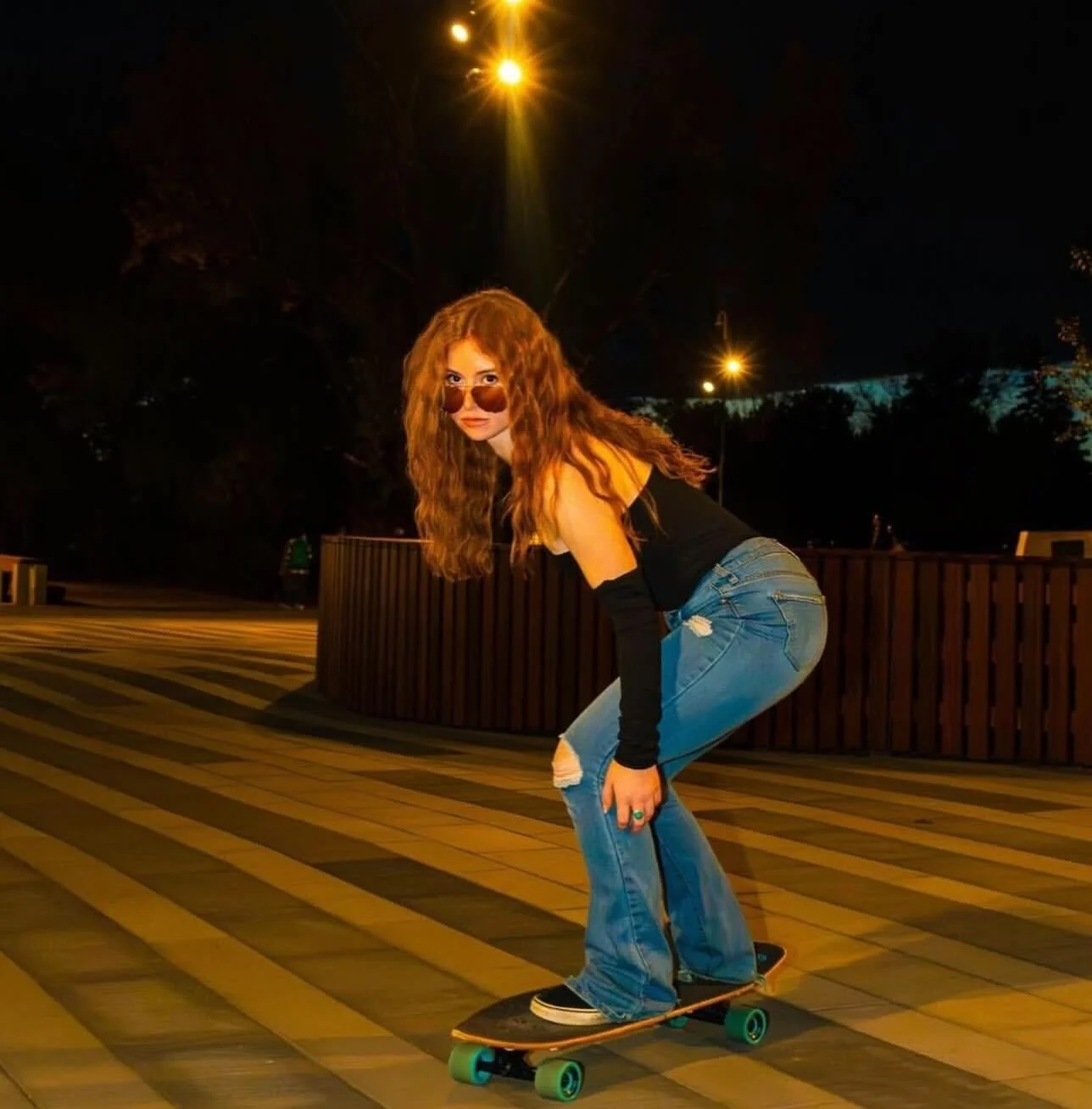 A young woman with long curly red hair, wearing sunglasses, a black sleeveless top, ripped jeans, and black shoes, skateboarding on a night street with streetlights and trees in the background.