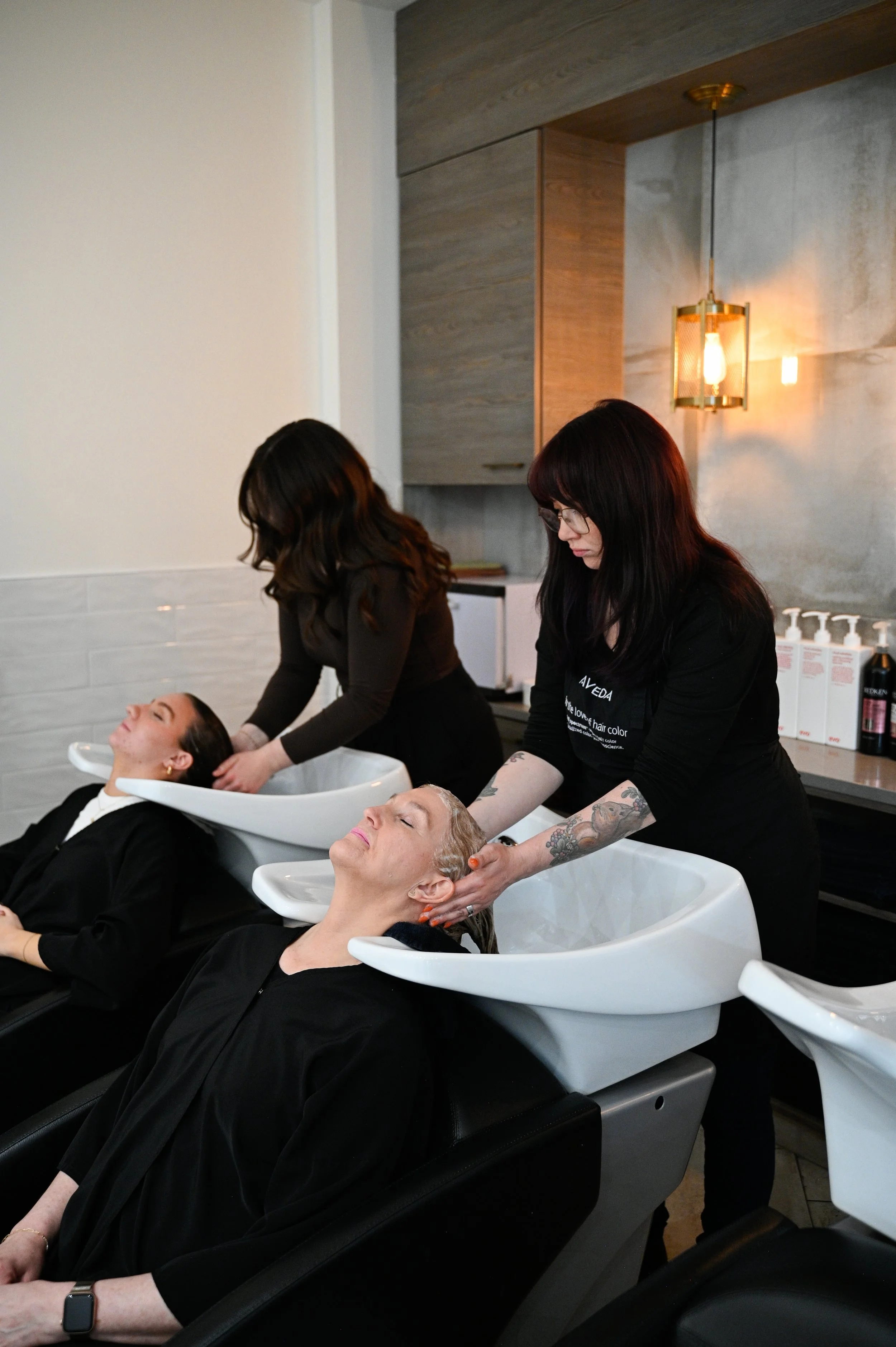Two women are reclining with their heads in hair washing sinks at a salon, while two stylists wash their hair collectively.