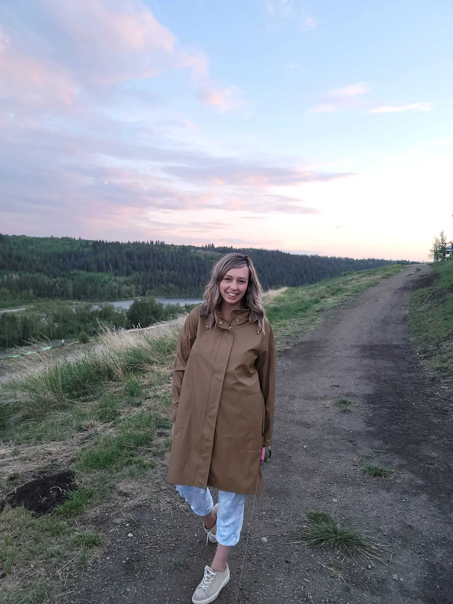 A woman standing on a dirt trail outdoors during sunset, wearing a tan coat, white pants, and sneakers, with a landscape of trees, a river, and a partly cloudy sky in the background.