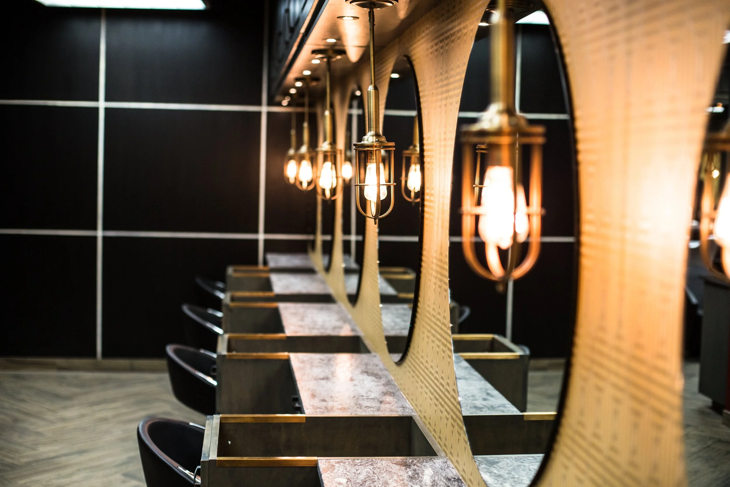 Interior of a modern hair salon or barbershop with marble-topped stations, black chairs, decorative mirrors, and vintage-style hanging light fixtures