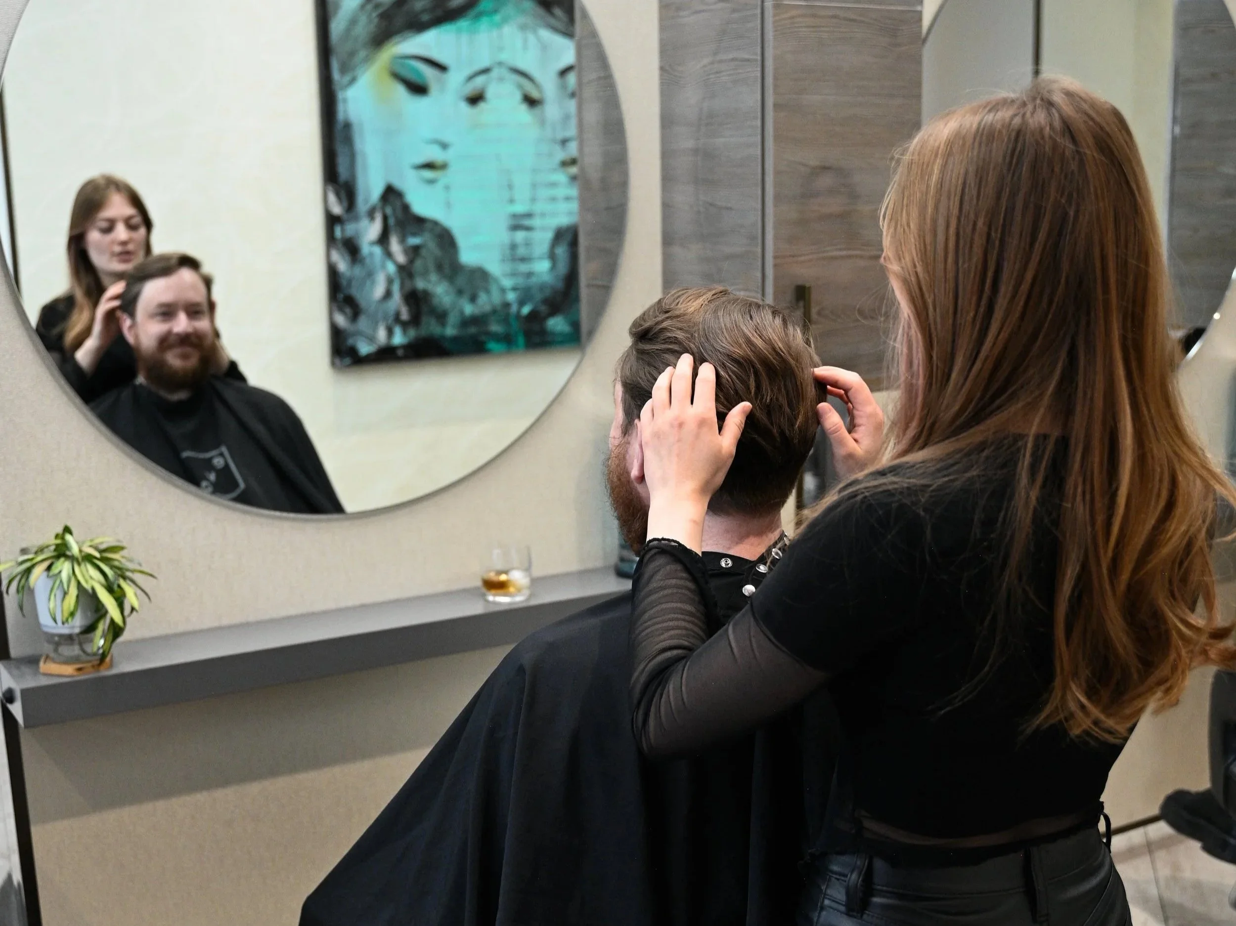 A woman with long red hair is giving a haircut to a man sitting in a salon chair, seen in a large round mirror. The salon has a small potted plant and a drink on a shelf. The man has brown hair and a beard, and the woman appears to be a hairstylist focusing on his hair.