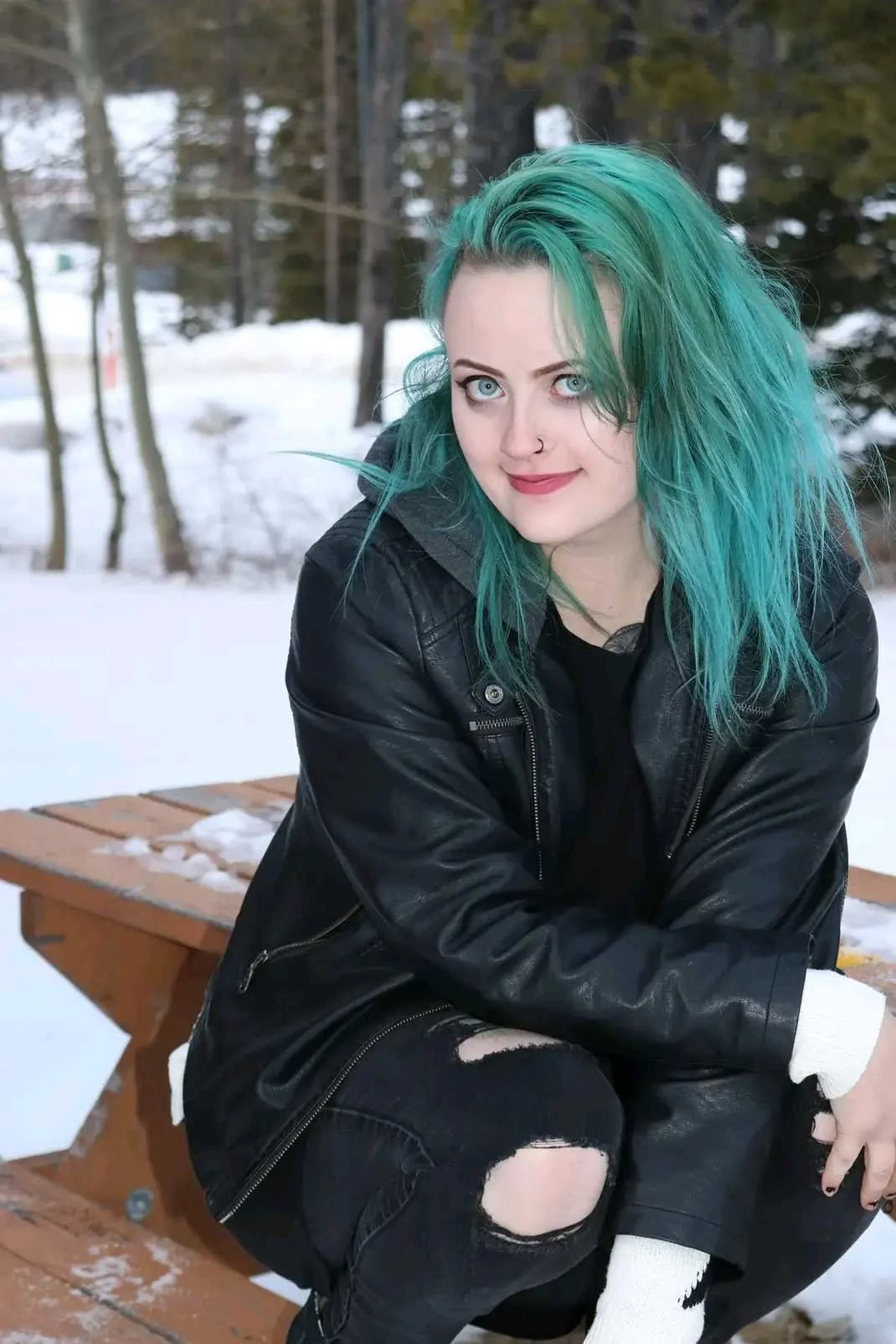 A young woman with bright turquoise hair, wearing a black leather jacket, sitting outdoors on a wooden bench in a snowy setting with trees in the background.