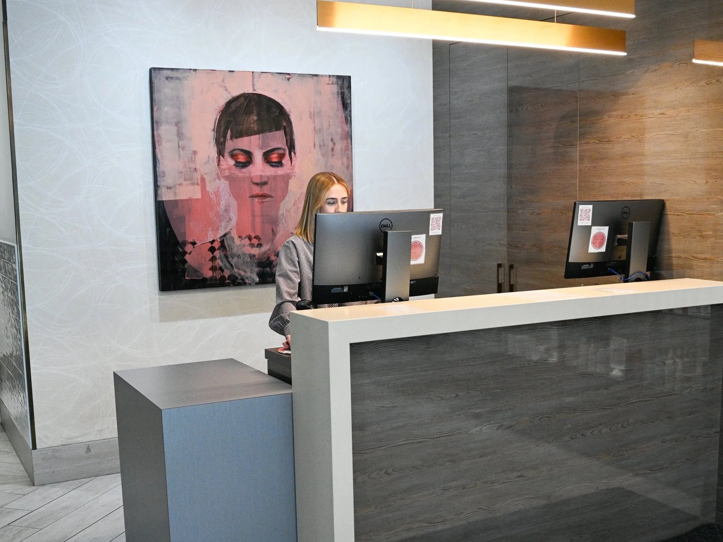 Reception area with a woman working on a computer behind a gray and white counter, a large framed portrait of a woman with short hair and closed eyes on the wall, and modern wooden and gray paneling