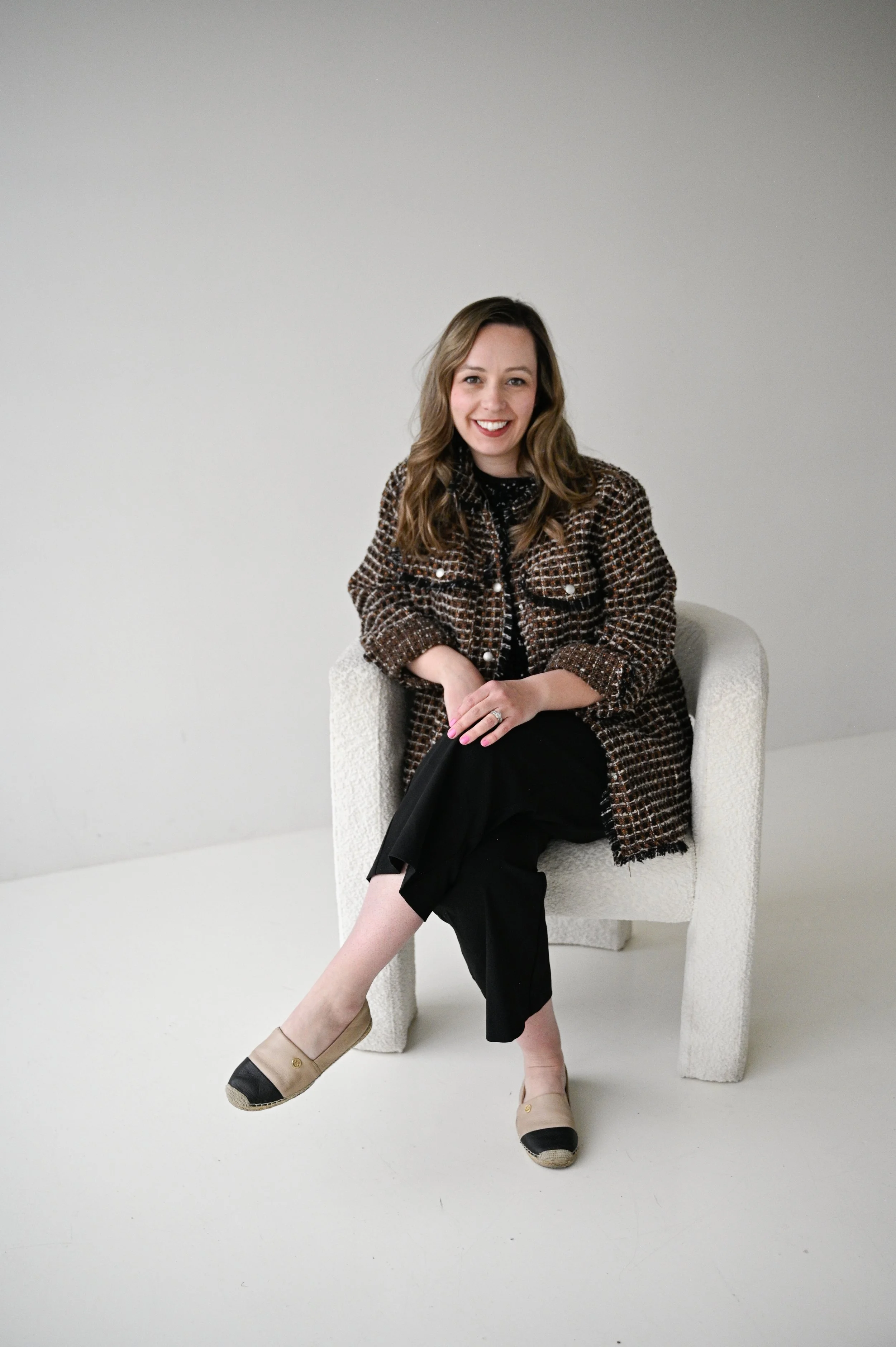 A woman with brown hair and a checkered jacket sitting on a white textured armchair, smiling, against a plain light gray wall.