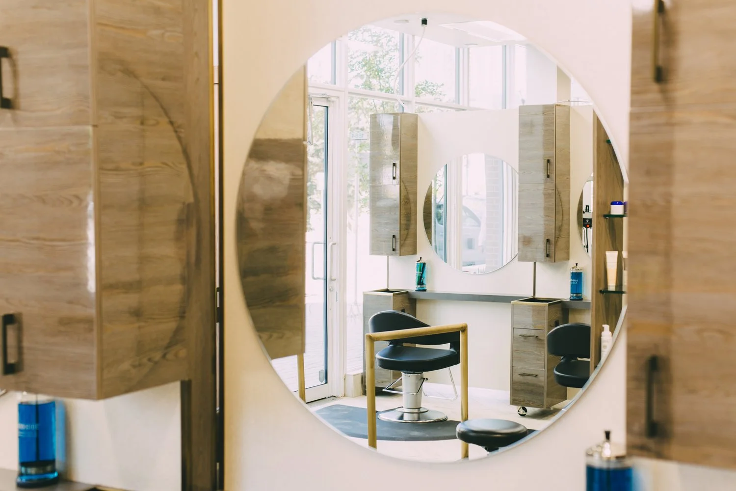 Salon view reflected in a large circular mirror showing a barber chair, wooden cabinets, and sunlight through large windows.
