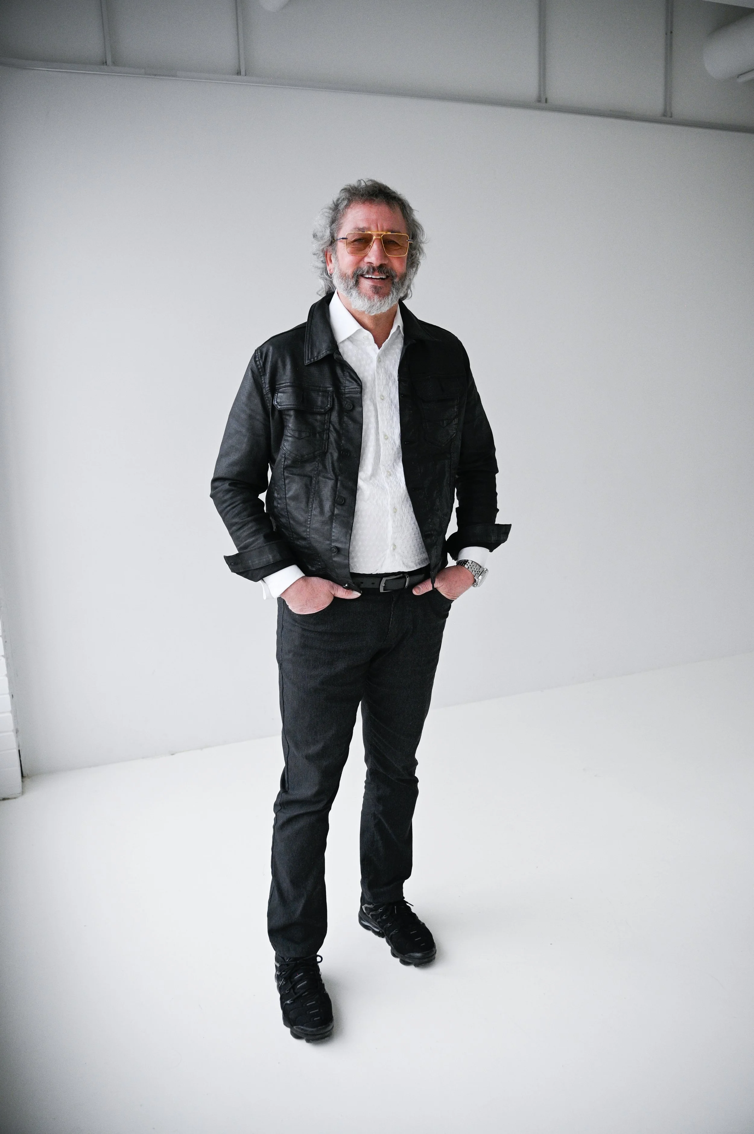 A man with gray hair, glasses, and a beard stands in a minimalistic space with white walls. He's wearing a black leather jacket, white shirt, dark pants, and black shoes, and has his hands in his pockets, smiling at the camera.