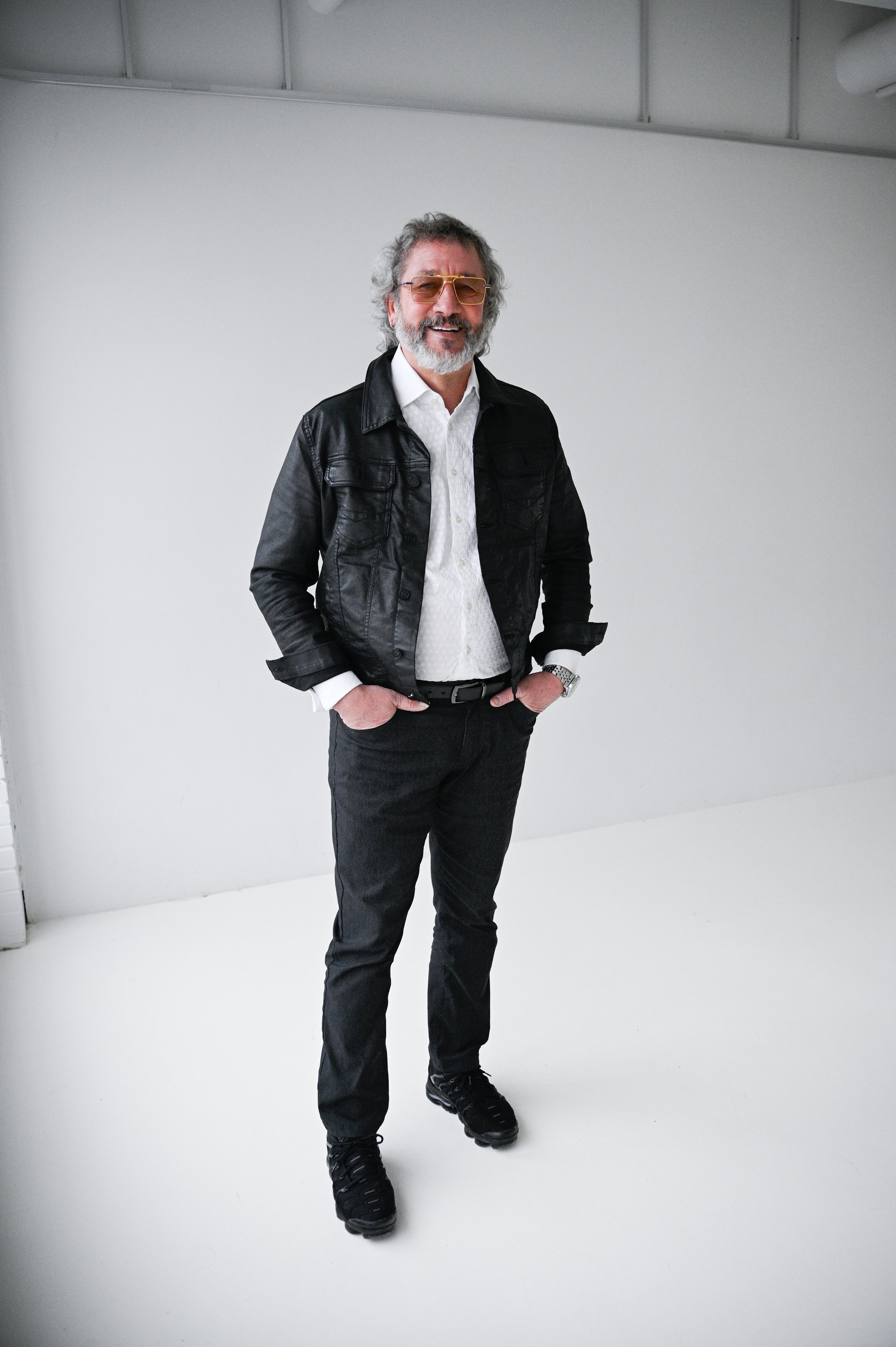 A man with gray hair and beard wearing sunglasses, a black leather jacket, white shirt, black pants, and black shoes, standing indoors against a plain white wall.