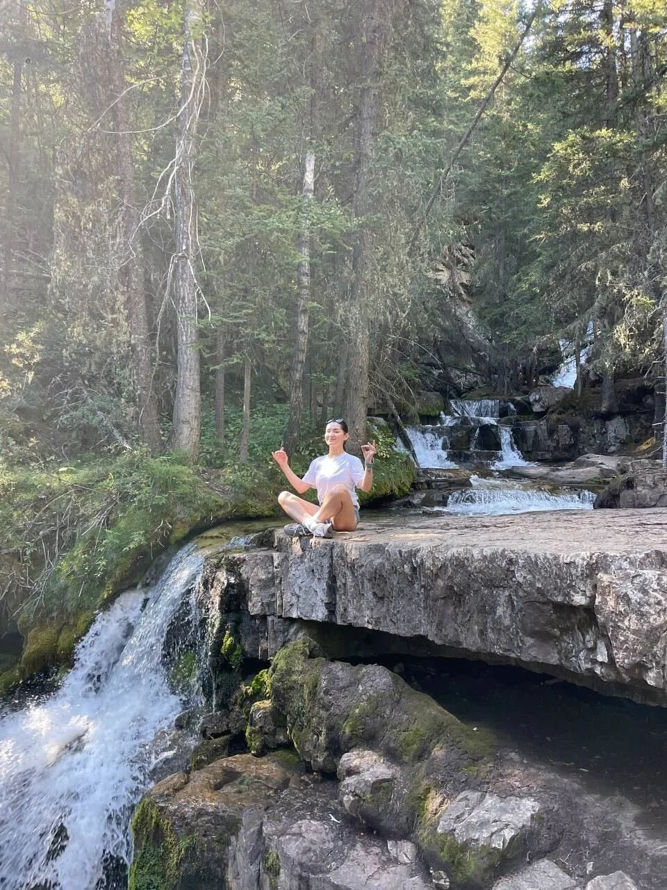 A person sitting cross-legged on a flat rock in a small waterfall, smiling and making hand gestures, surrounded by a forest with tall trees and cascading water.