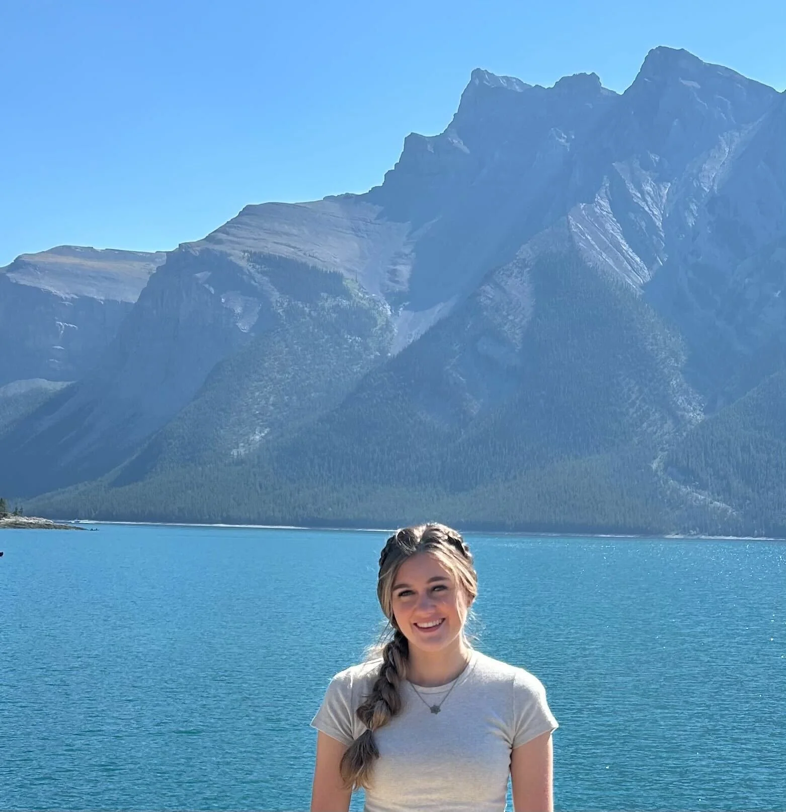 A young woman with a braid and white t-shirt smiling in front of a large blue lake with mountains and a clear blue sky in the background.