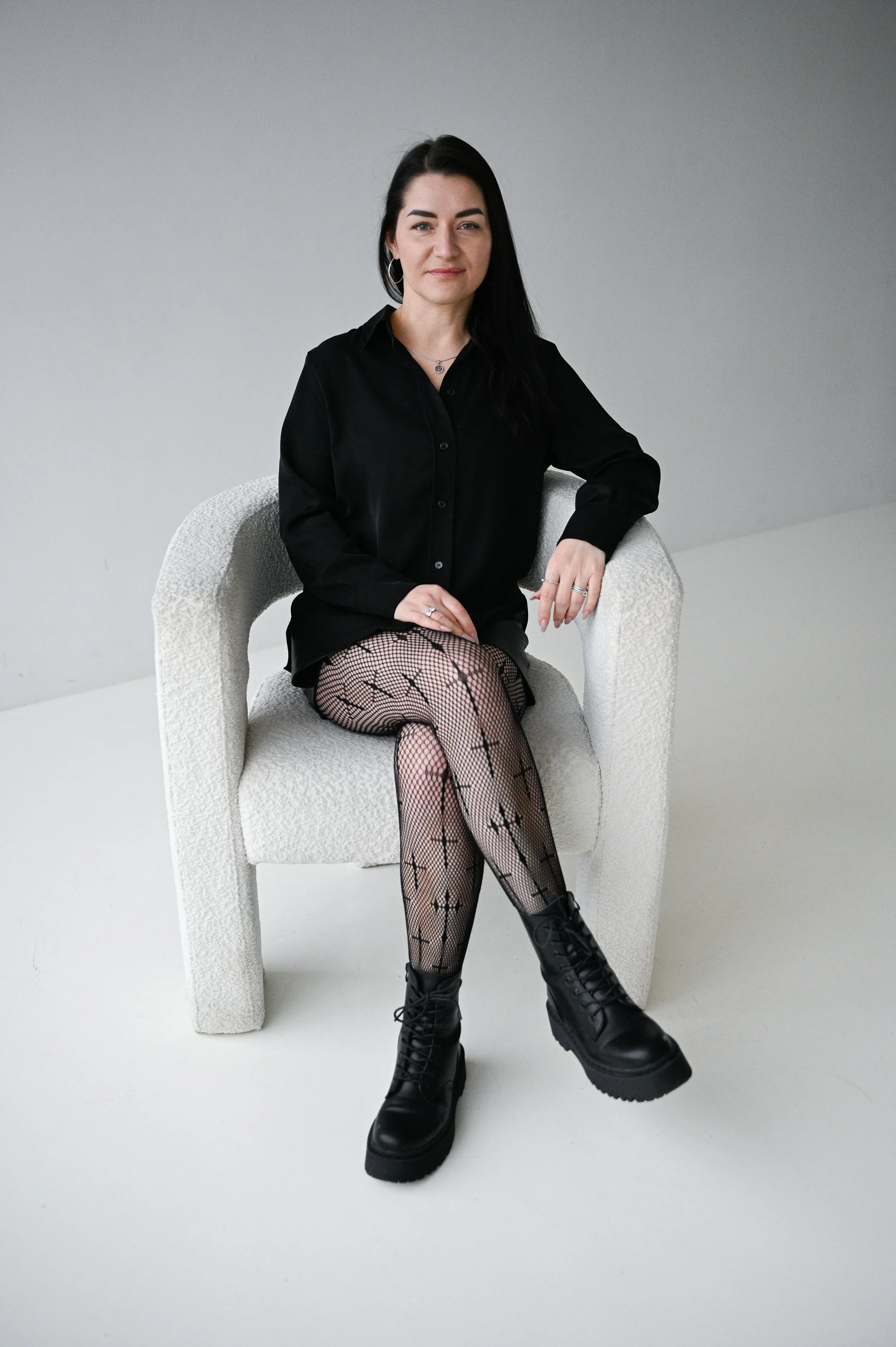 A woman sitting on a light-colored armchair in a minimalist setting, wearing a black shirt, black fishnet tights with crosses, and black combat boots.