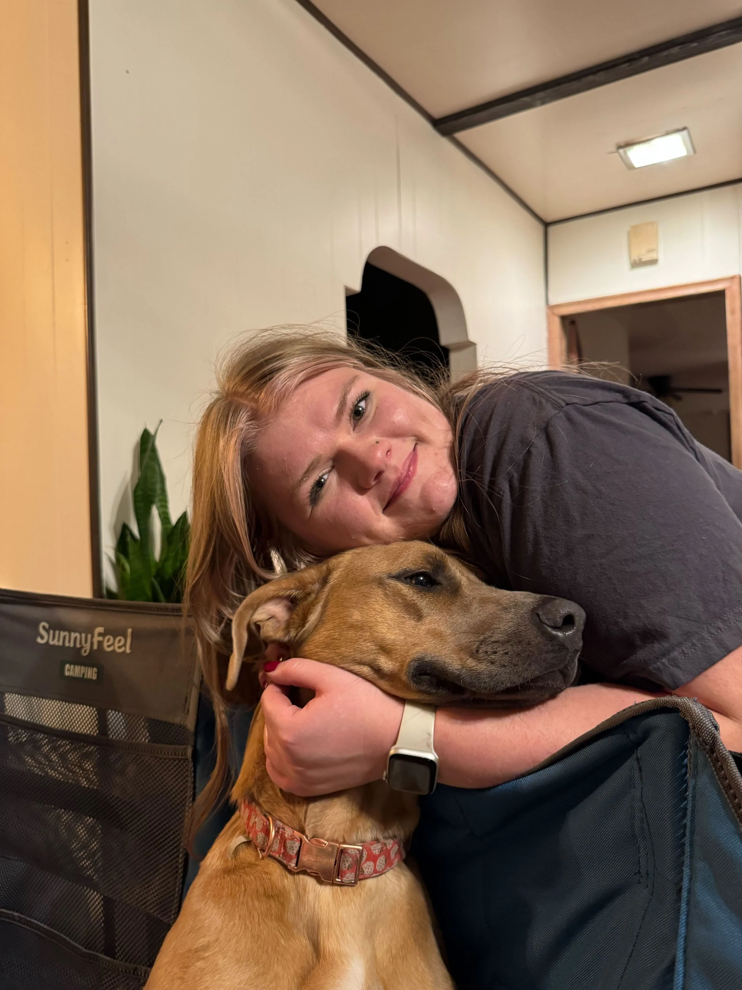 A woman hugging a large brown dog lying on a backpack in an indoor room, smiling at the camera.
