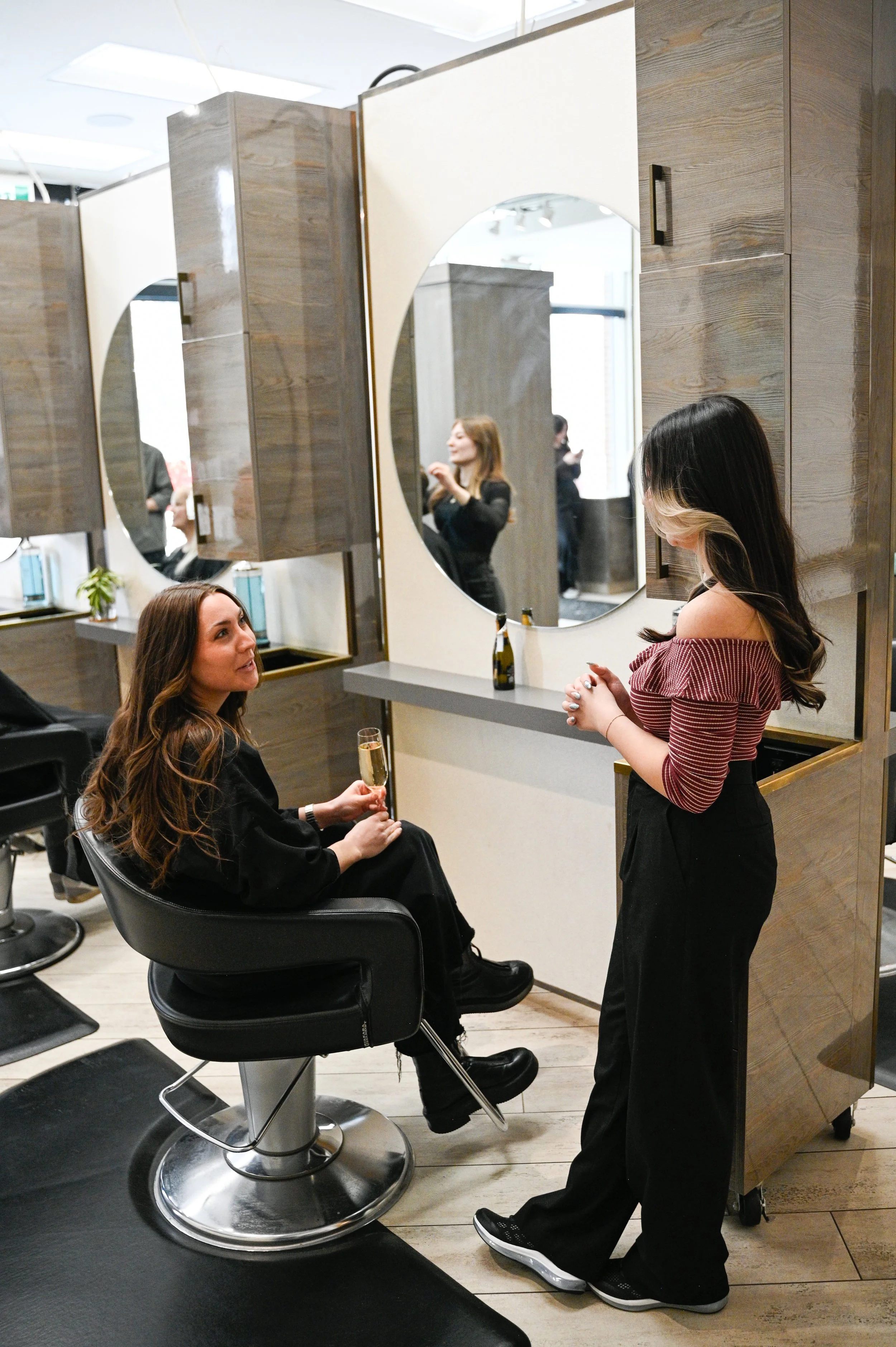 A woman with long brown hair, sitting in a black salon chair, holding a glass of champagne, and talking to a hairstylist with long black hair and blonde highlights. They are in a modern salon with wood cabinets and large mirrors.