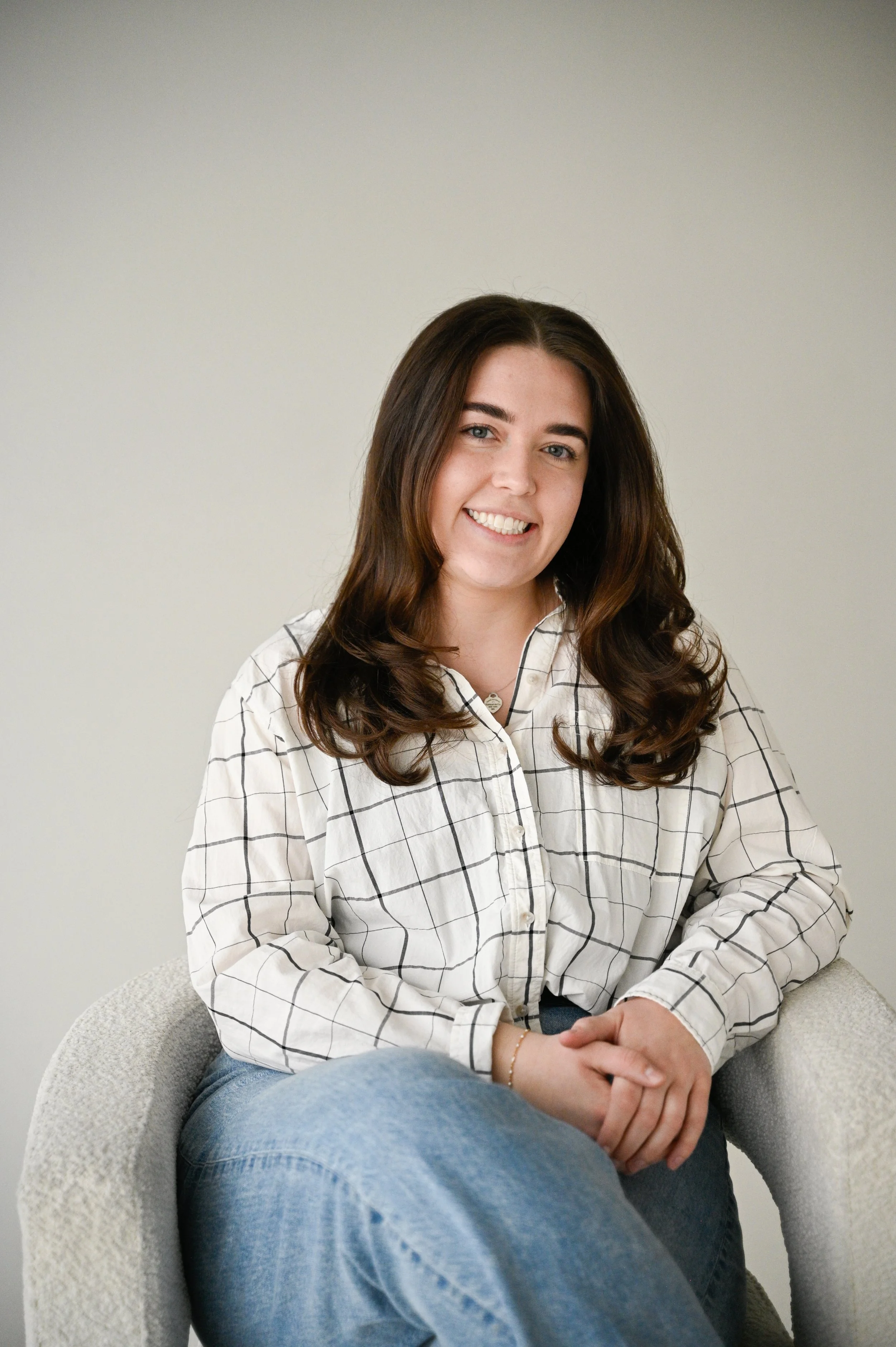 A young woman with long, wavy brown hair sitting in a light-colored armchair against a plain wall. She is smiling and wearing a white button-up shirt with black grid lines and blue jeans.