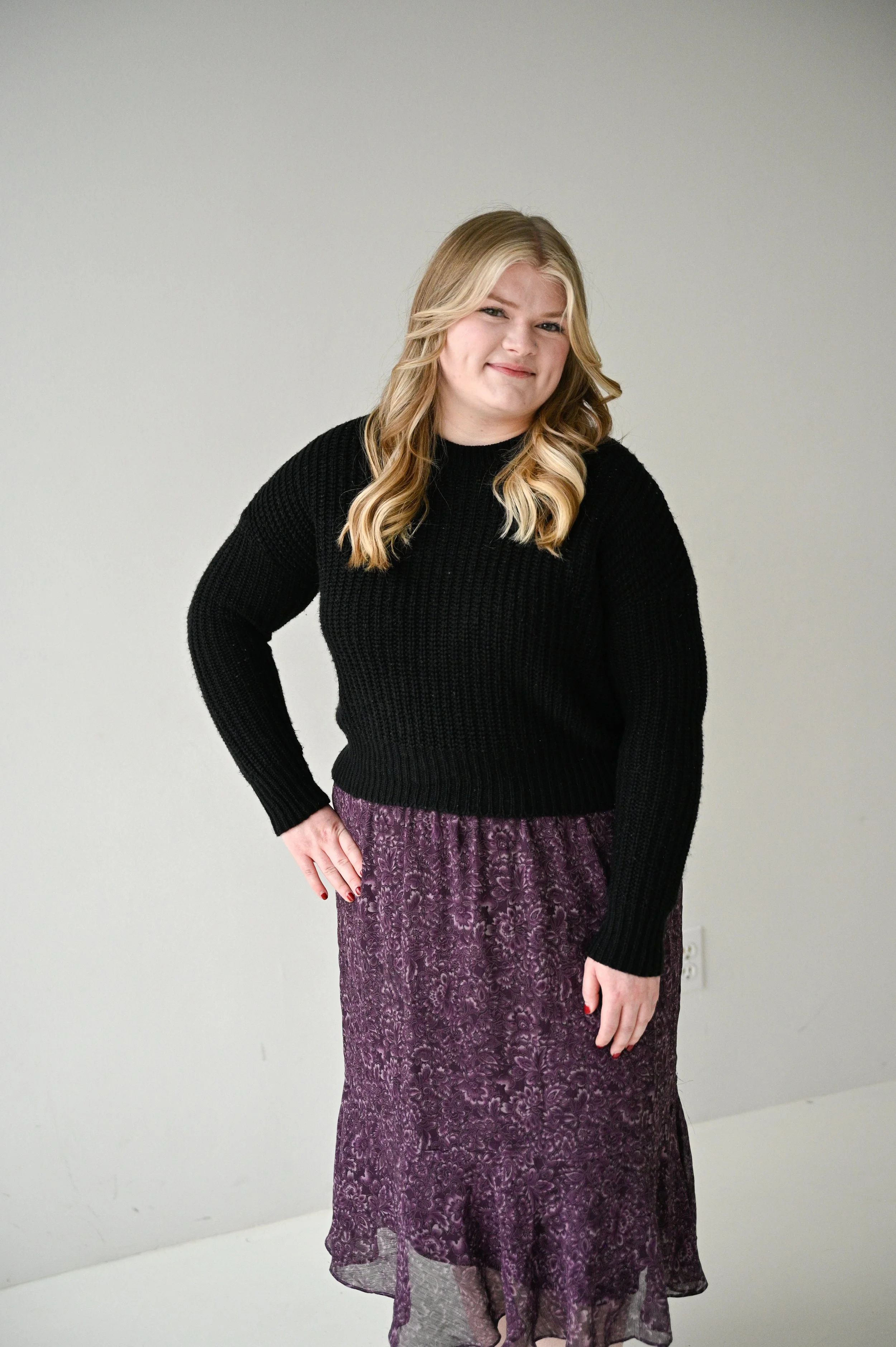 A woman with blonde hair winks at the camera, wearing a black sweater and a purple lace skirt, standing against a plain light-colored background.