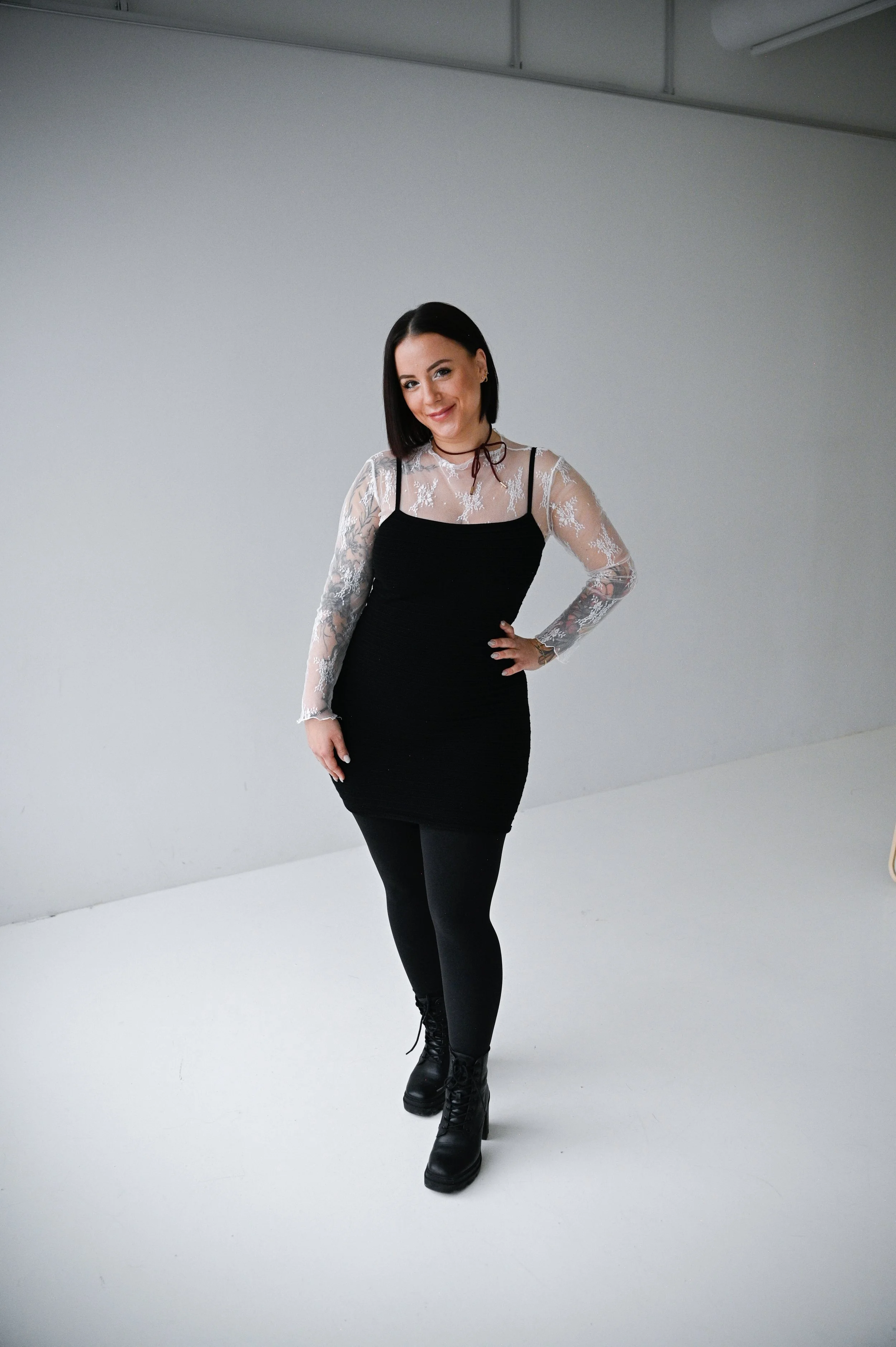 A woman with shoulder-length black hair, wearing a black dress with sheer lace long sleeves and a lace top, black leggings, and black boots, standing in front of a plain white background.