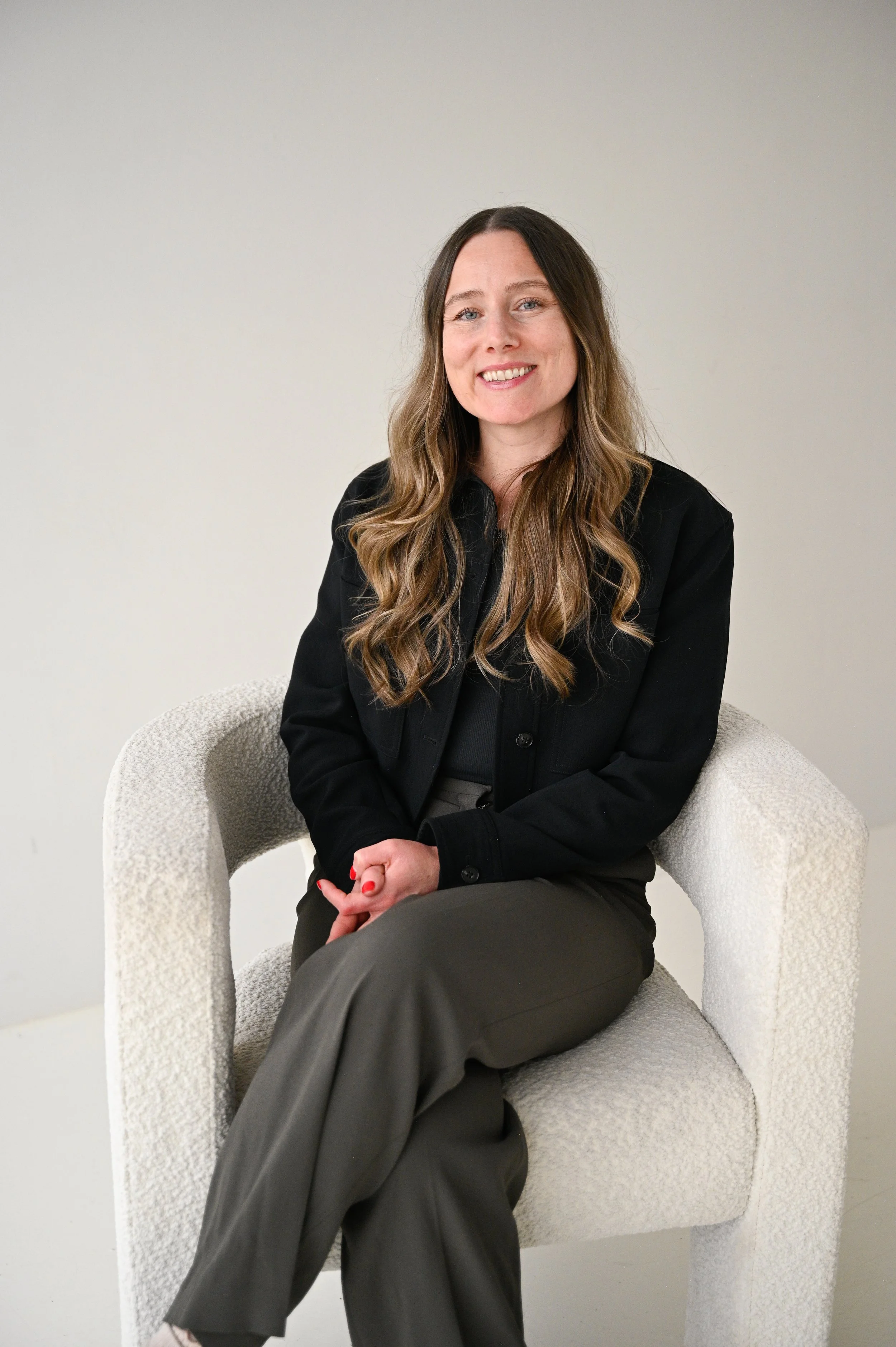 A woman with long wavy hair, dressed in a black jacket and gray pants, sitting on a light-colored textured armchair against a plain white wall, smiling at the camera.