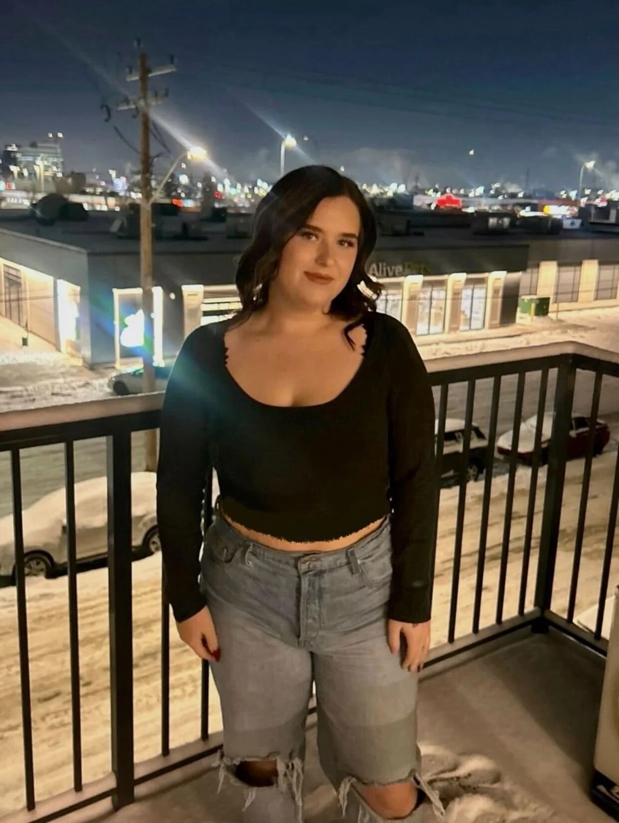 A young woman with dark hair and a black top, standing on a balcony at night, overlooking a snowy parking lot and commercial street with lit storefronts and cars.