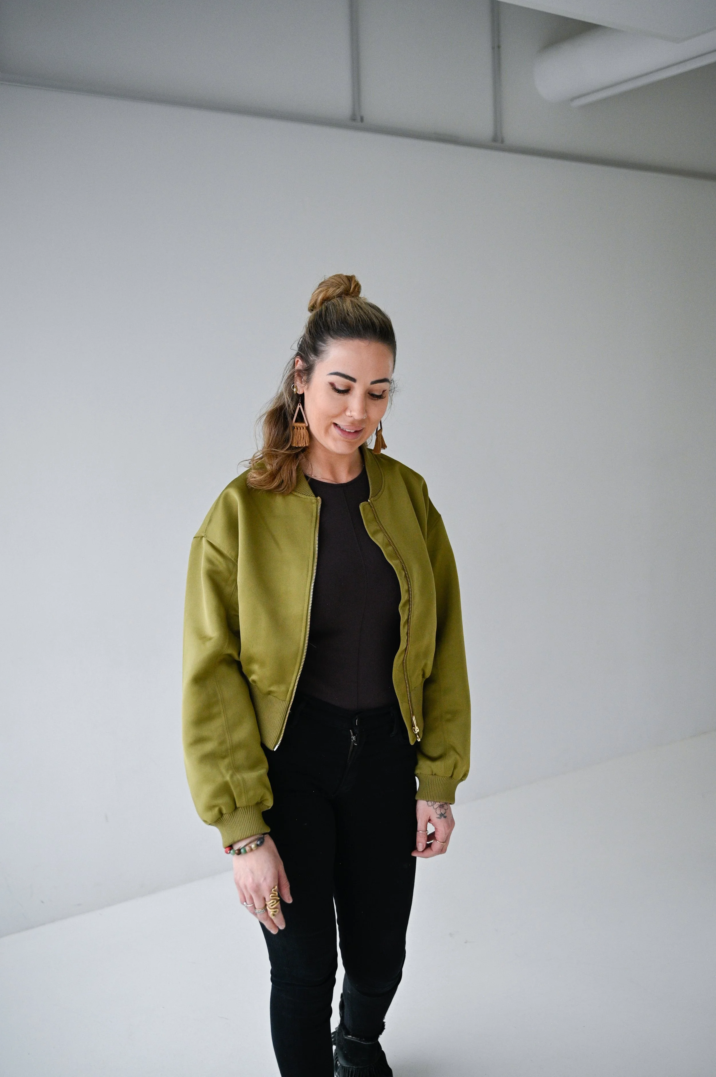 A woman with long brown hair in a messy bun, wearing gold statement earrings, a black top, black pants, and an olive green bomber jacket, looking down with a slight smile.