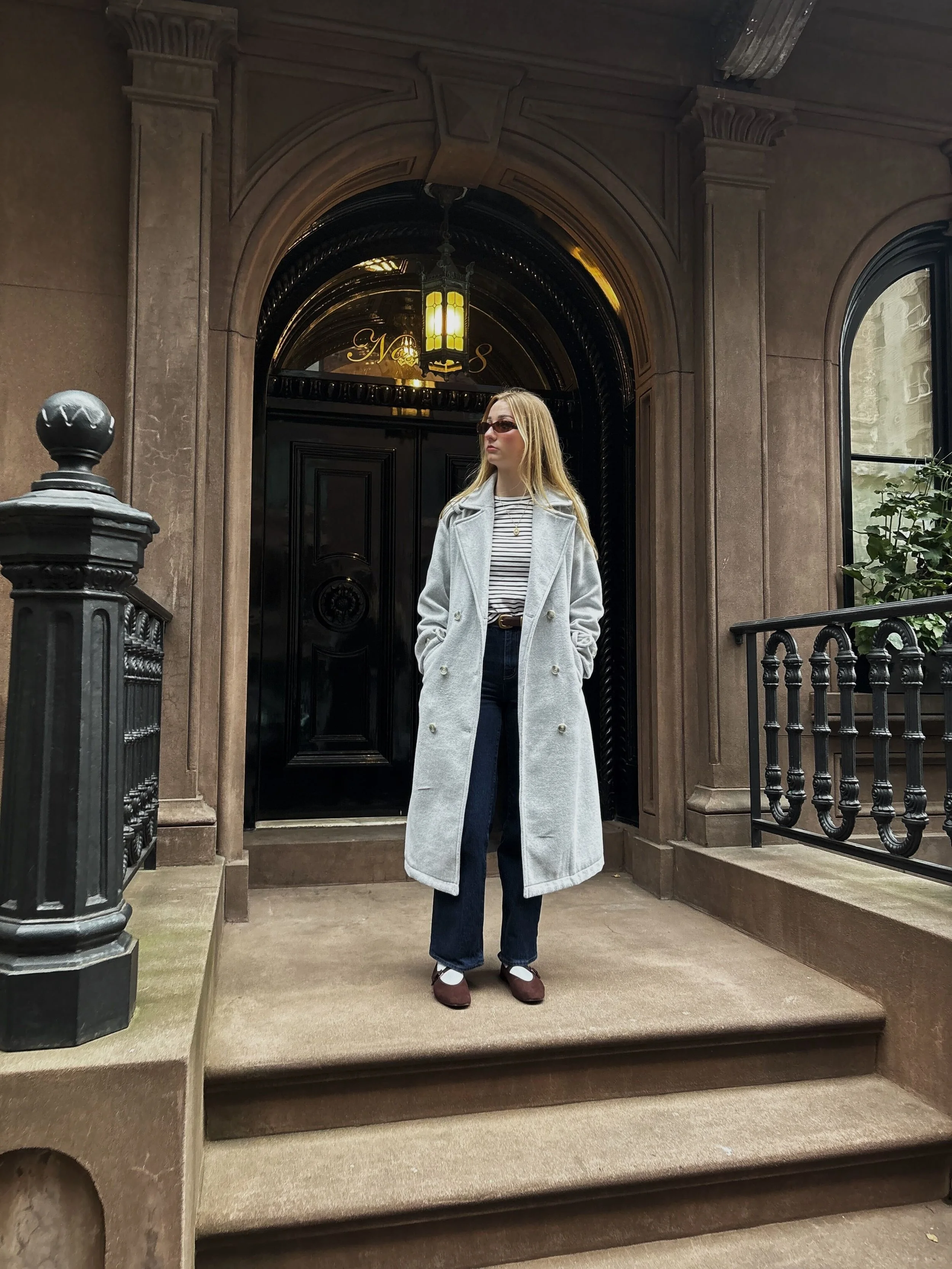 A woman with blonde hair wearing sunglasses, a striped shirt, dark jeans, brown shoes, and a light gray coat is standing on a city sidewalk in front of a black door with gold accents, under an arched entryway.