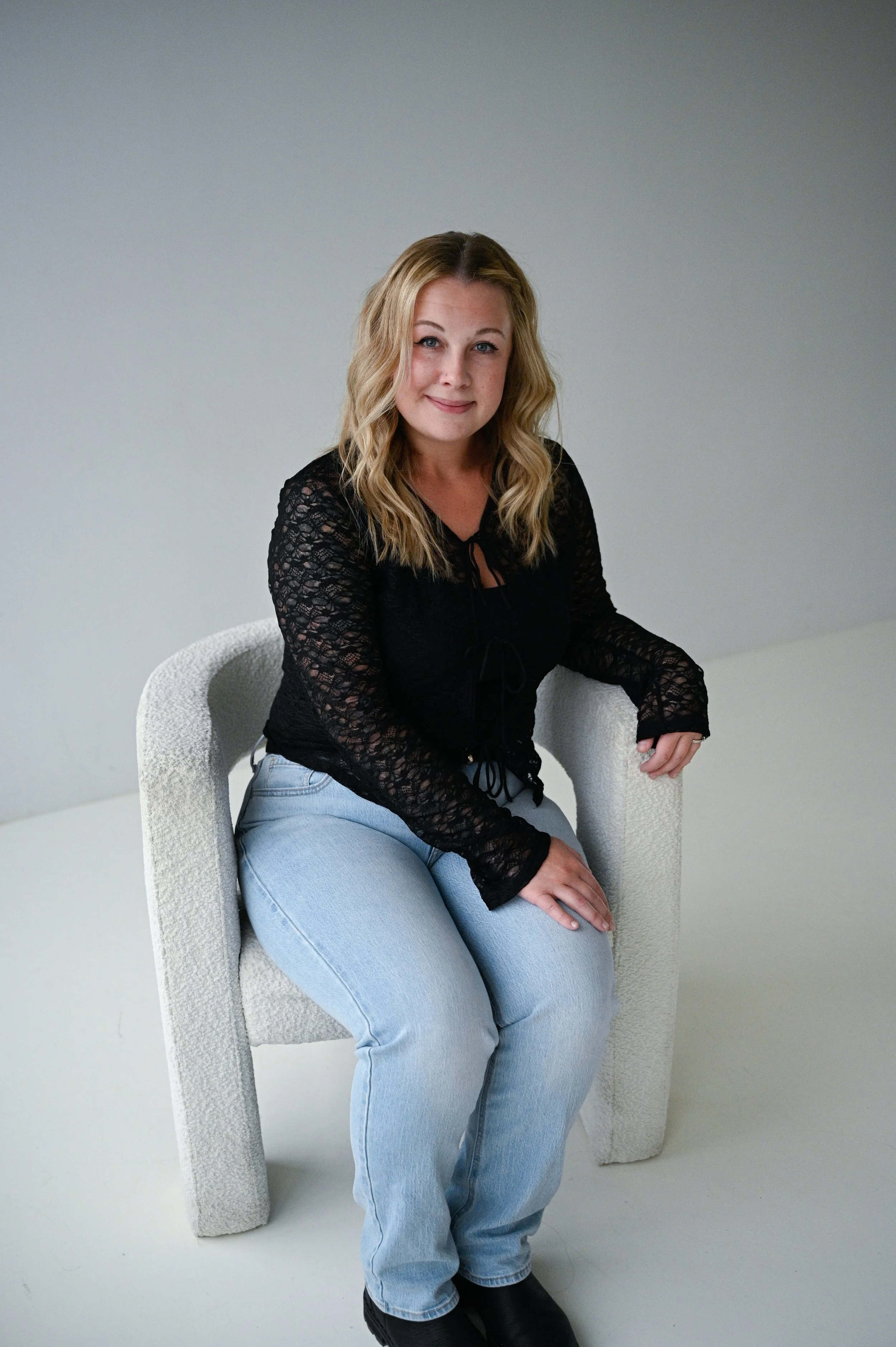 A woman with blonde wavy hair sitting on a light-colored textured chair, wearing a black lace long sleeve top and light blue jeans, in a plain light gray room.