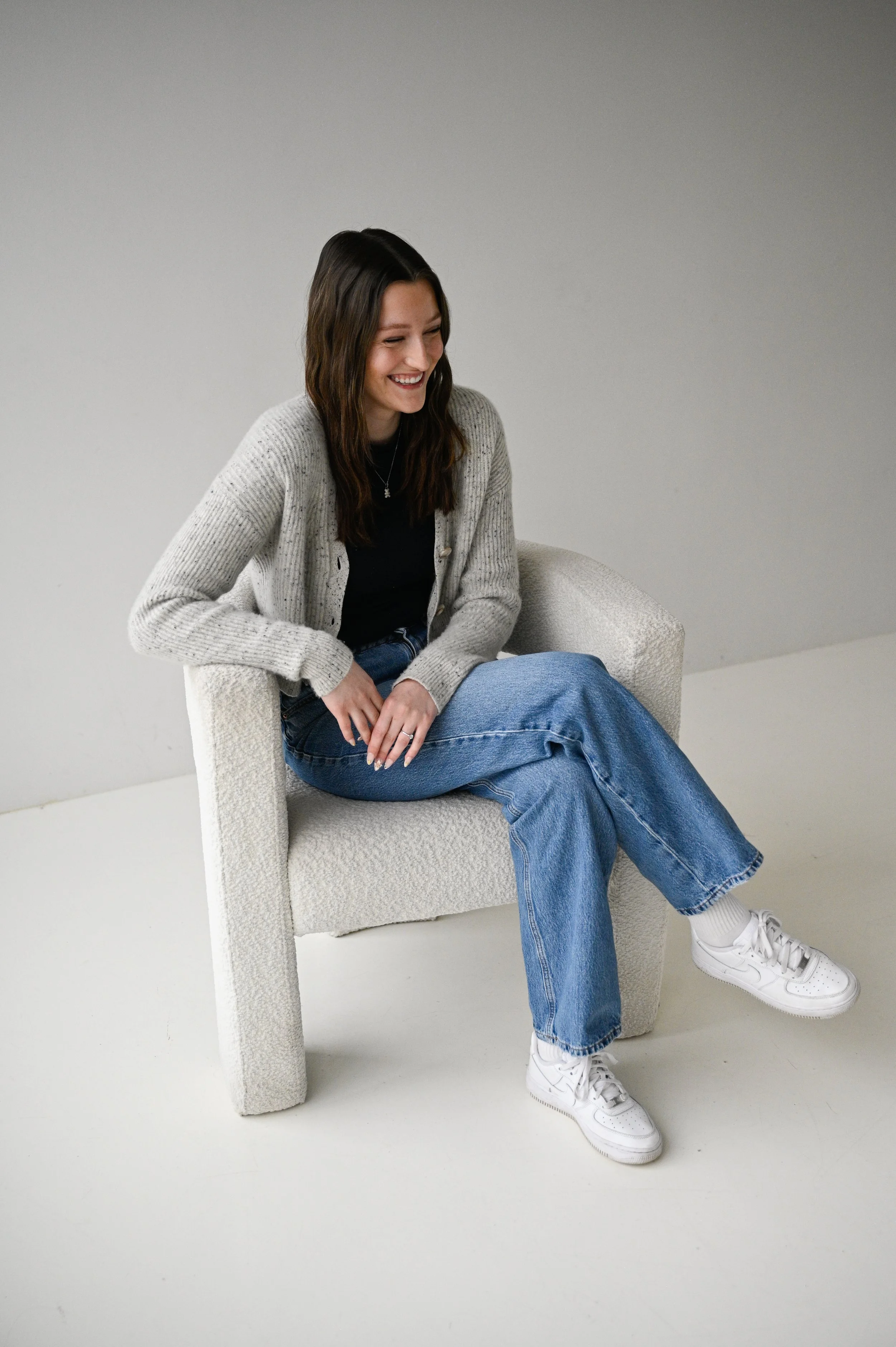 A young woman with brown hair laughing, sitting on a light-colored armchair in a plain, light gray room, dressed casually in a black shirt, gray cardigan, blue jeans, and white sneakers.