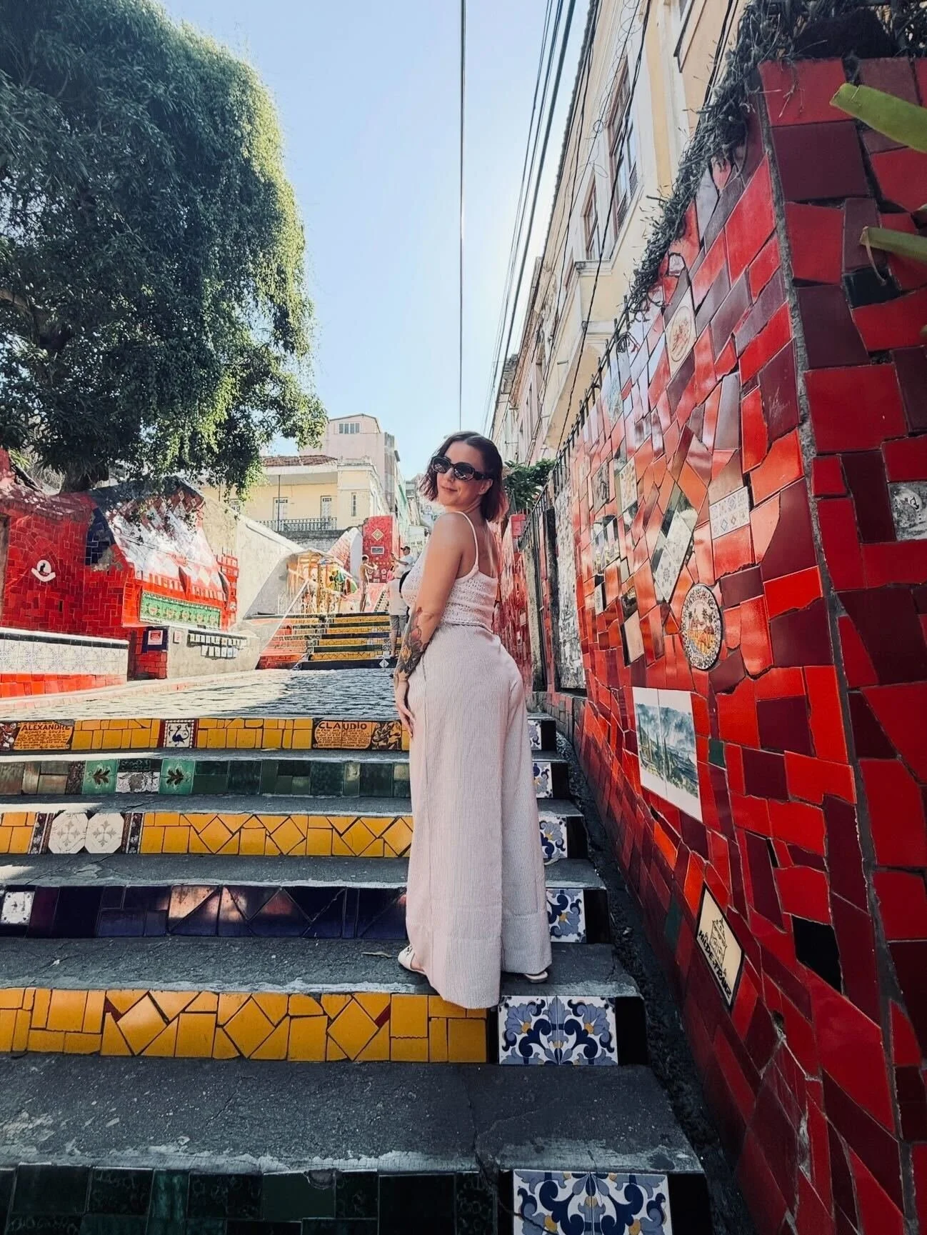 A woman in white pants and sunglasses standing on colorful tiled stairs next to a red mosaic wall, with buildings and a tree in the background.