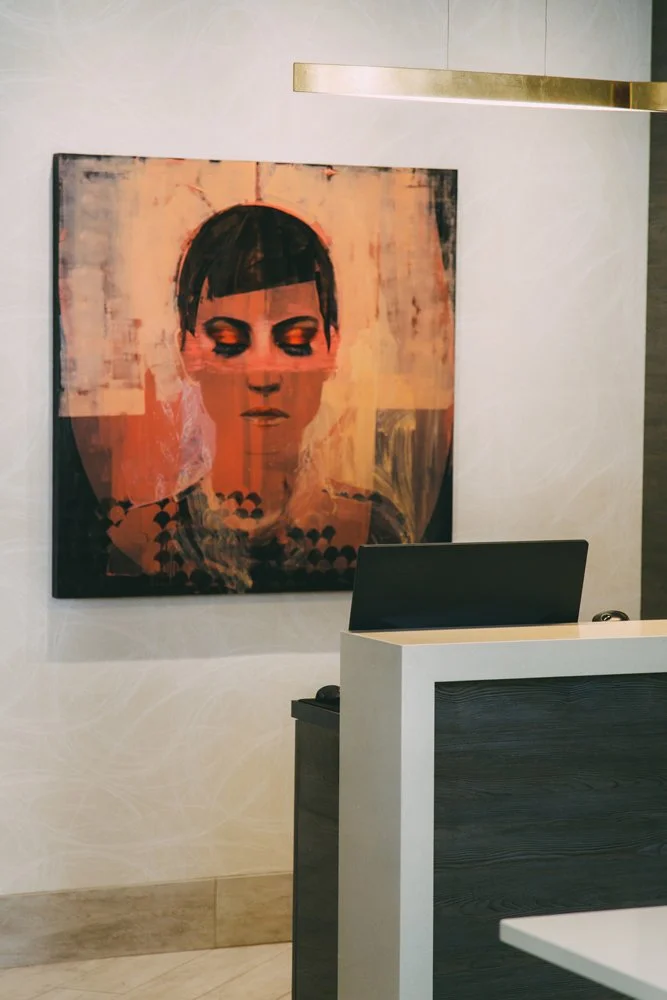 A colorful abstract portrait painting of a woman with short dark hair, closed eyes, and makeup, mounted on a white wall in a modern interior space.