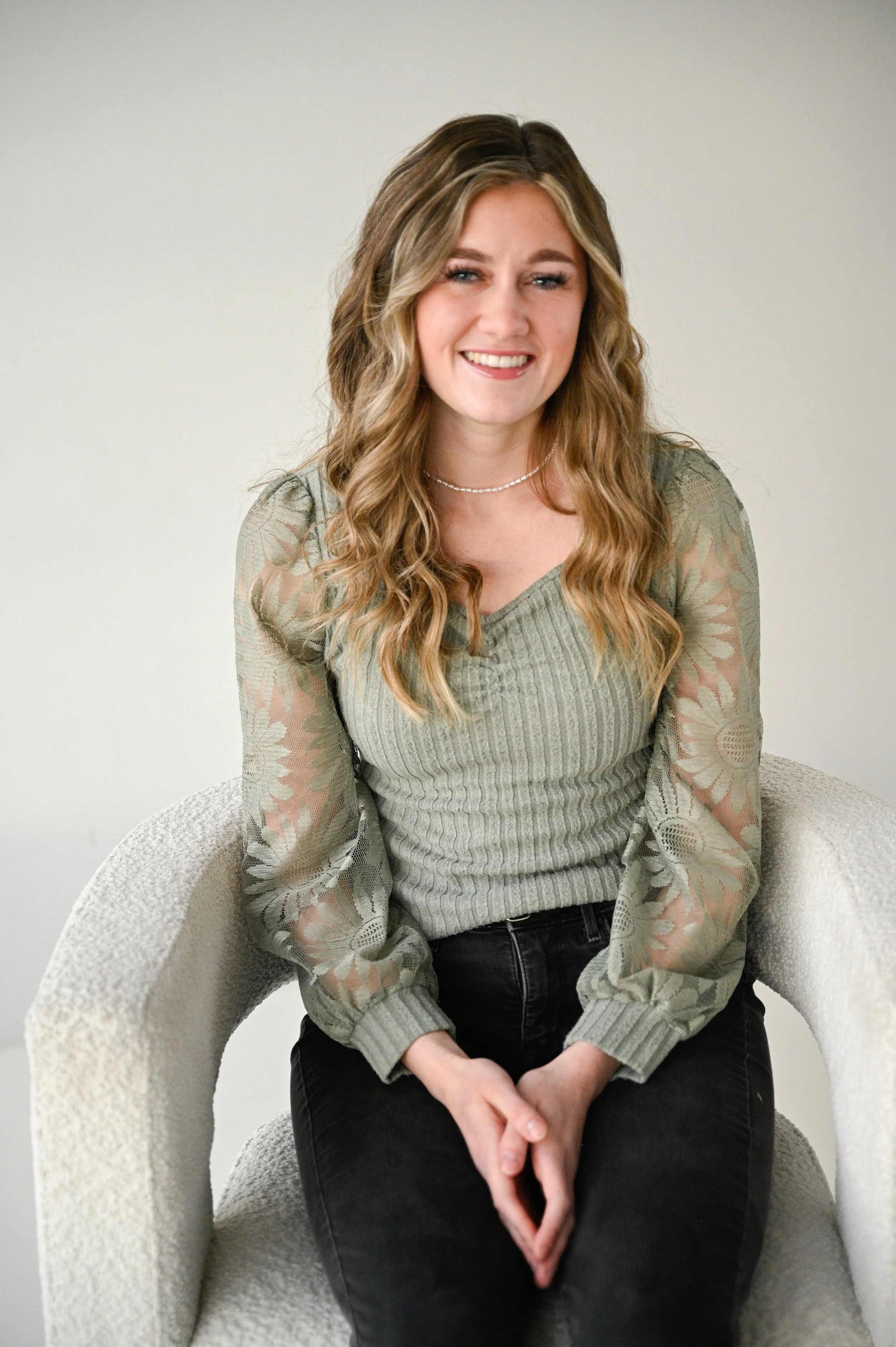 A young woman with long wavy blonde hair, wearing a green long-sleeved blouse with floral lace sleeves and black pants, sitting on a light-colored textured armchair, smiling at the camera.