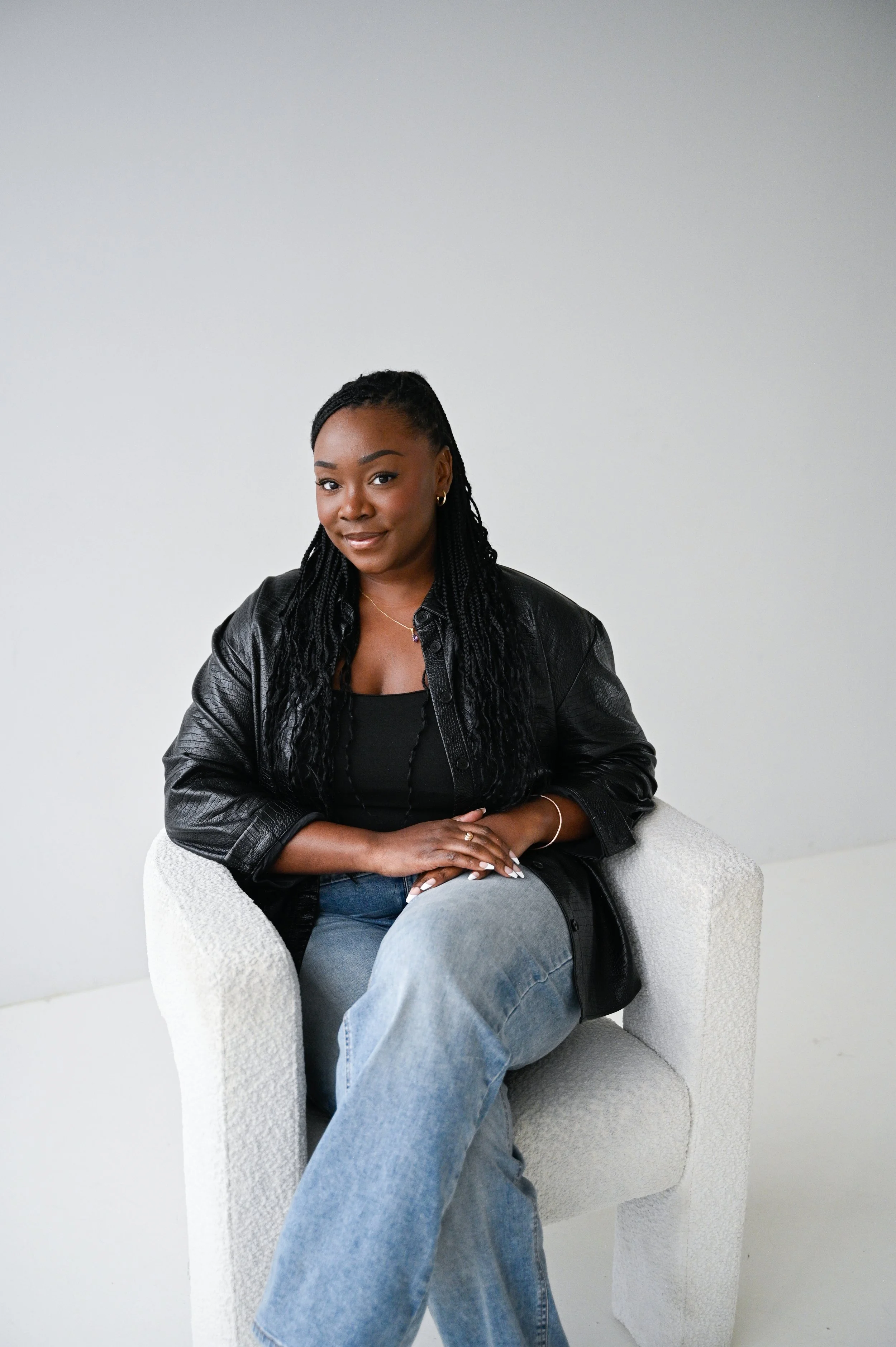 A woman with dark braided hair sits in a white armchair against a plain white wall, wearing a black leather jacket and blue jeans.